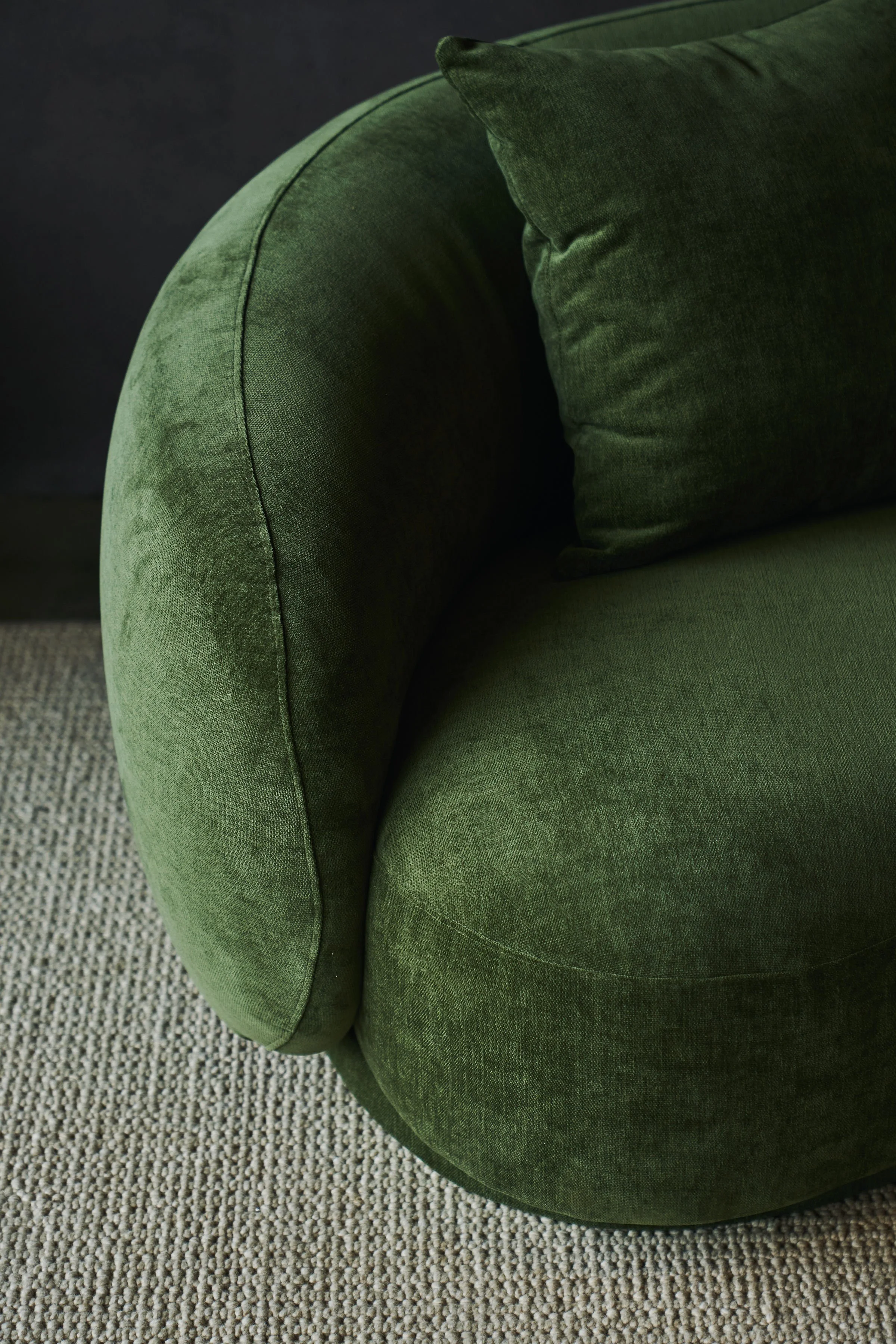 Close-up of a green upholstered armchair with a matching green cushion, placed on a textured beige carpet with a dark background.