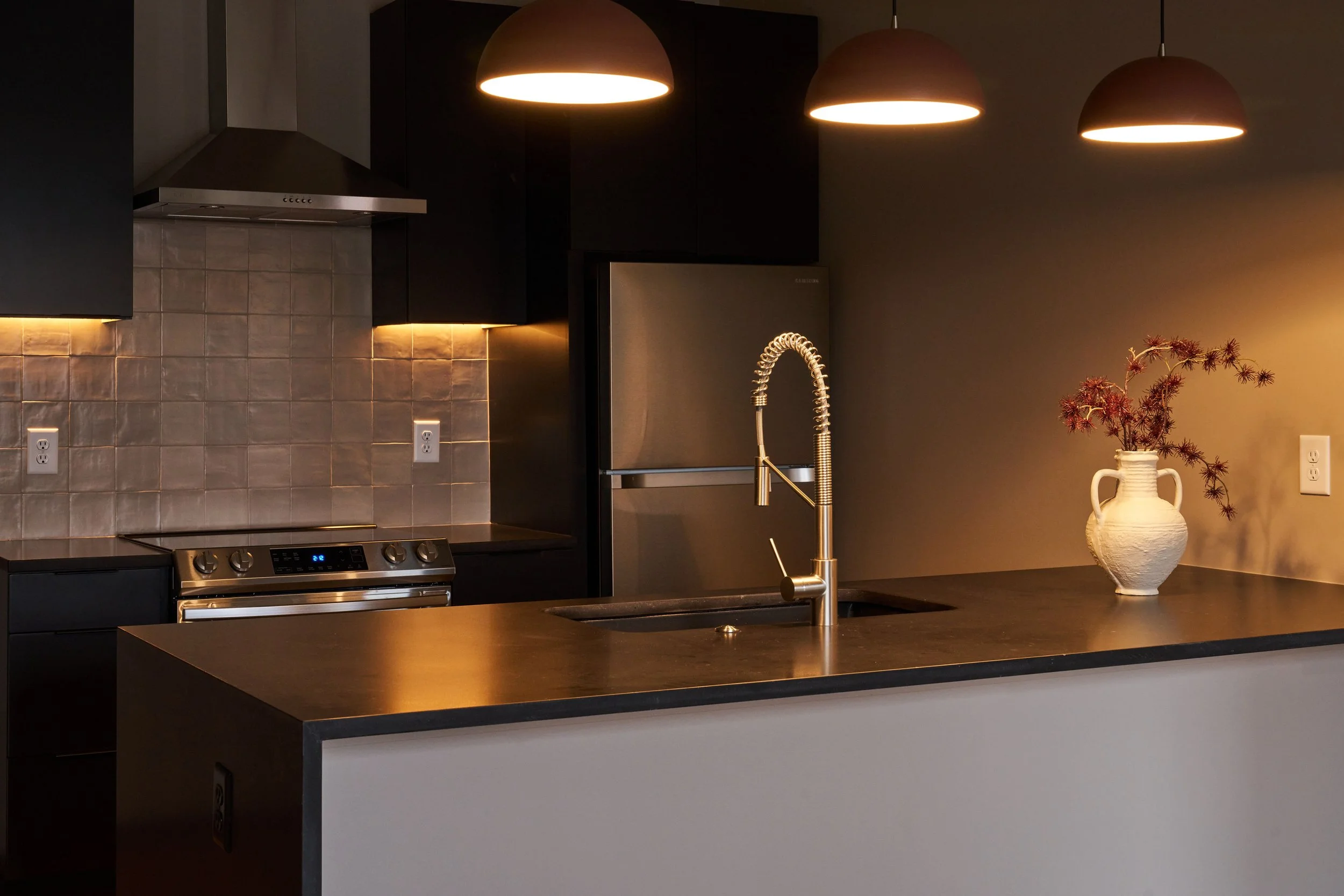 Modern kitchen with black cabinets, industrial style pendant lighting, a metallic stovetop, a stainless steel refrigerator, a black countertop, a white vase with dried flowers, and warm under-cabinet lighting.