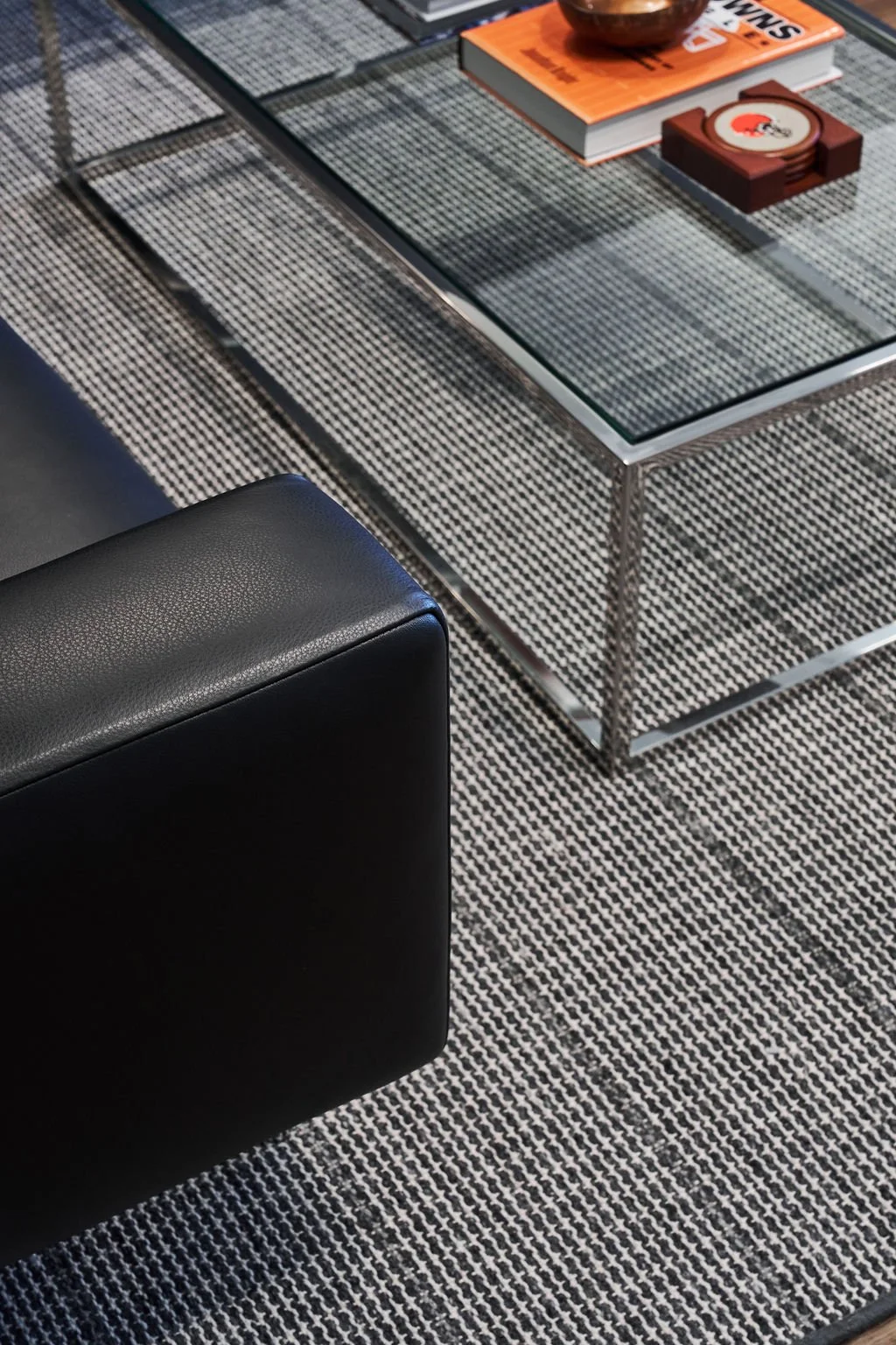 A glass-top coffee table with magazines and a coaster on a black-and-white woven rug, next to a black leather ottoman.