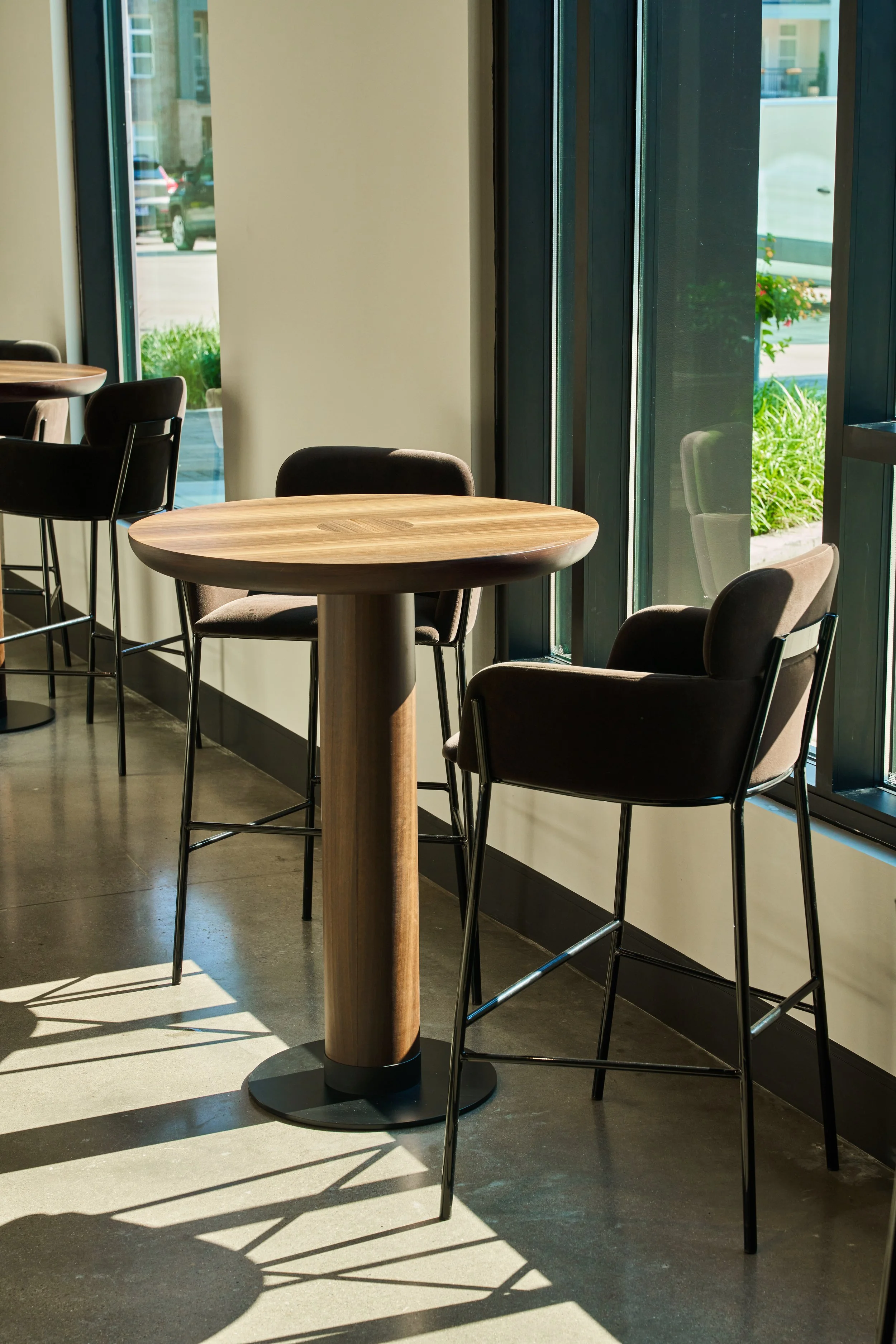 Interior of a cafe or restaurant with wooden tables, black chairs, large windows letting in sunlight, and outdoor greenery visible through the windows.