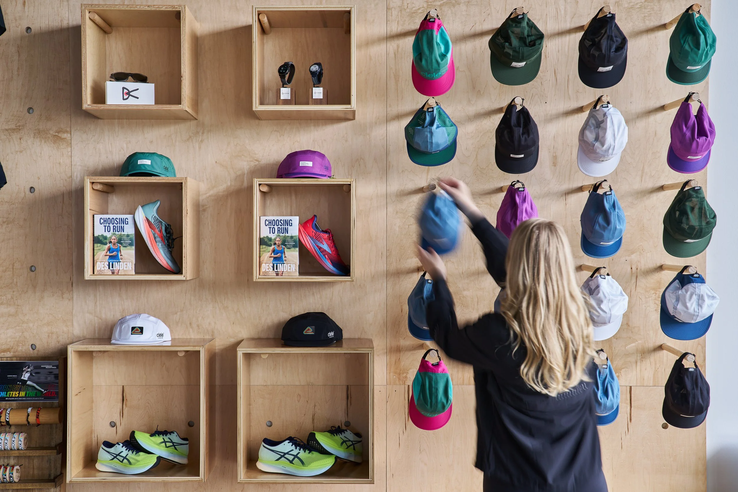 A person with long blonde hair shopping for hats and visors at a sports store. The display wall is made of light-colored wood with hats hanging on hooks in various colors, including pink, green, black, blue, purple, and white. There are also shelves 