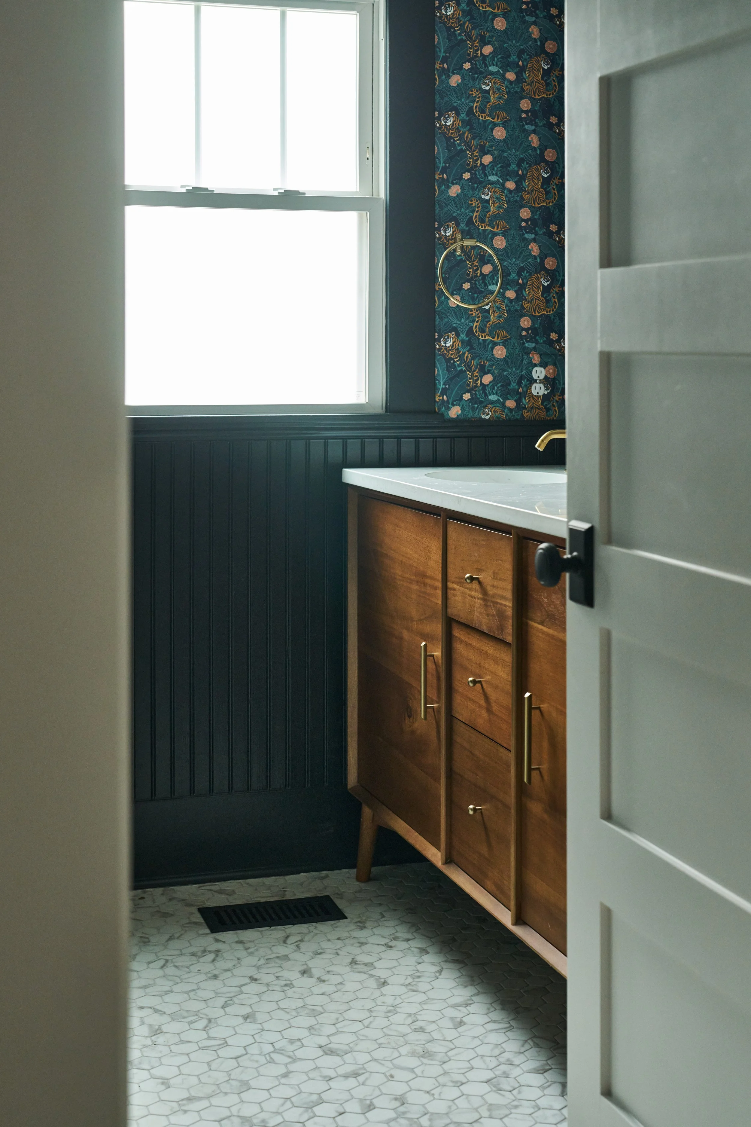 A bathroom with a window, a dark blue wainscoting, a floral wallpaper with tigers, a round towel ring, a double power outlet, a wooden vanity with gold handles, a white marble countertop with an integrated sink, and hexagonal white floor tiles.