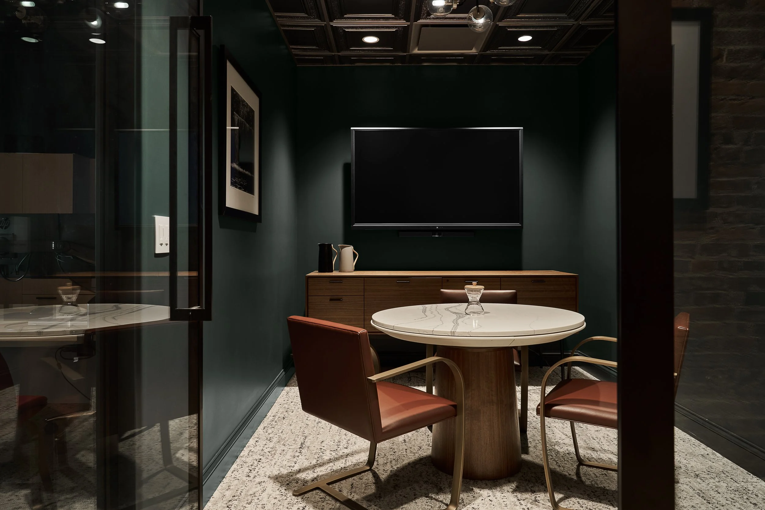 A modern, dimly-lit dining area with a round marble table, four chairs with red cushions, a wooden sideboard with decorative vases, a large empty flat-screen TV on a green wall, and framed artwork, all viewed from outside through a glass door.