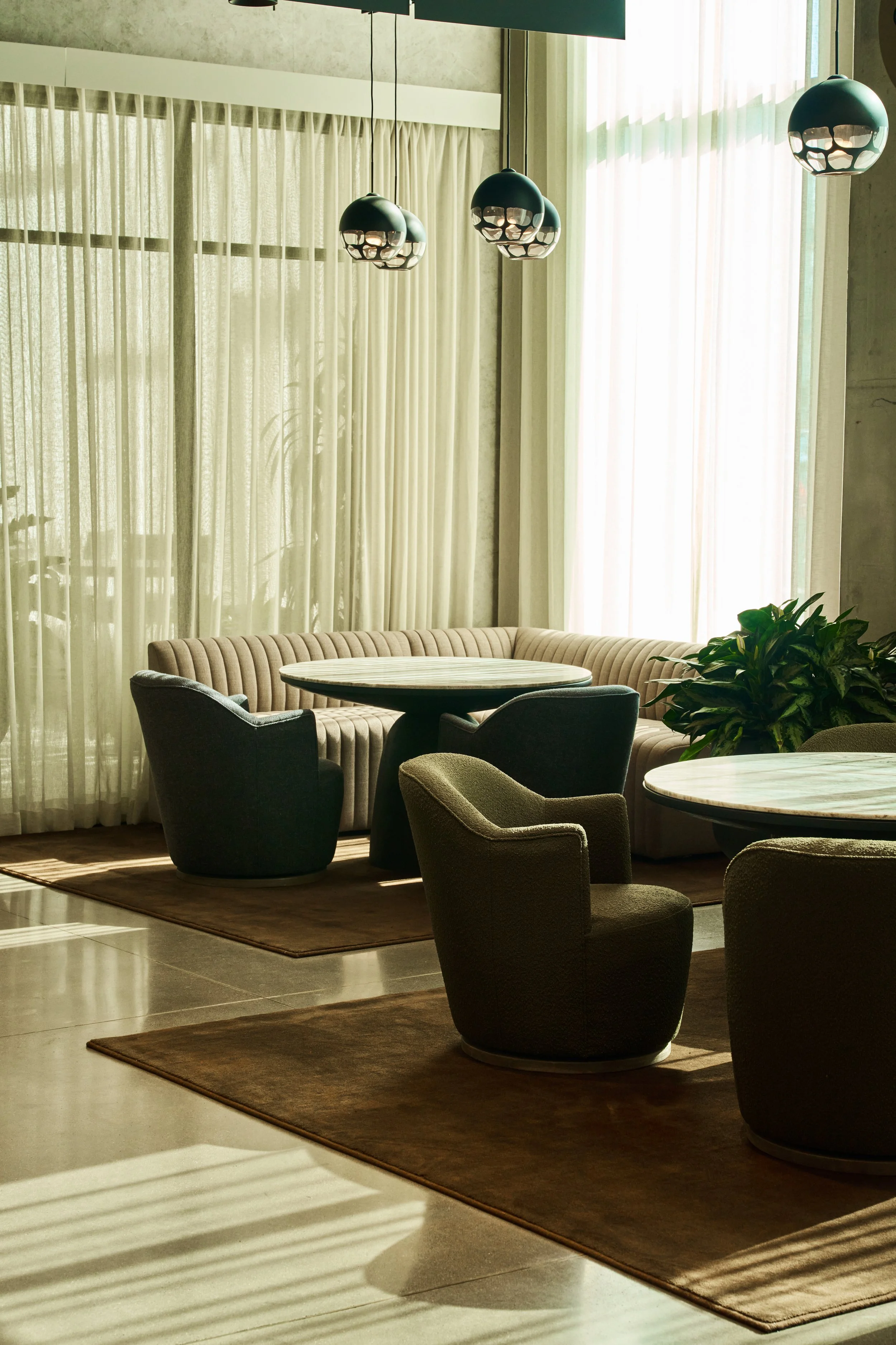 Interior of a modern restaurant or lounge with round marble-top tables, upholstered chairs, large windows with sheer curtains, hanging pendant lights, and a plant in the corner.