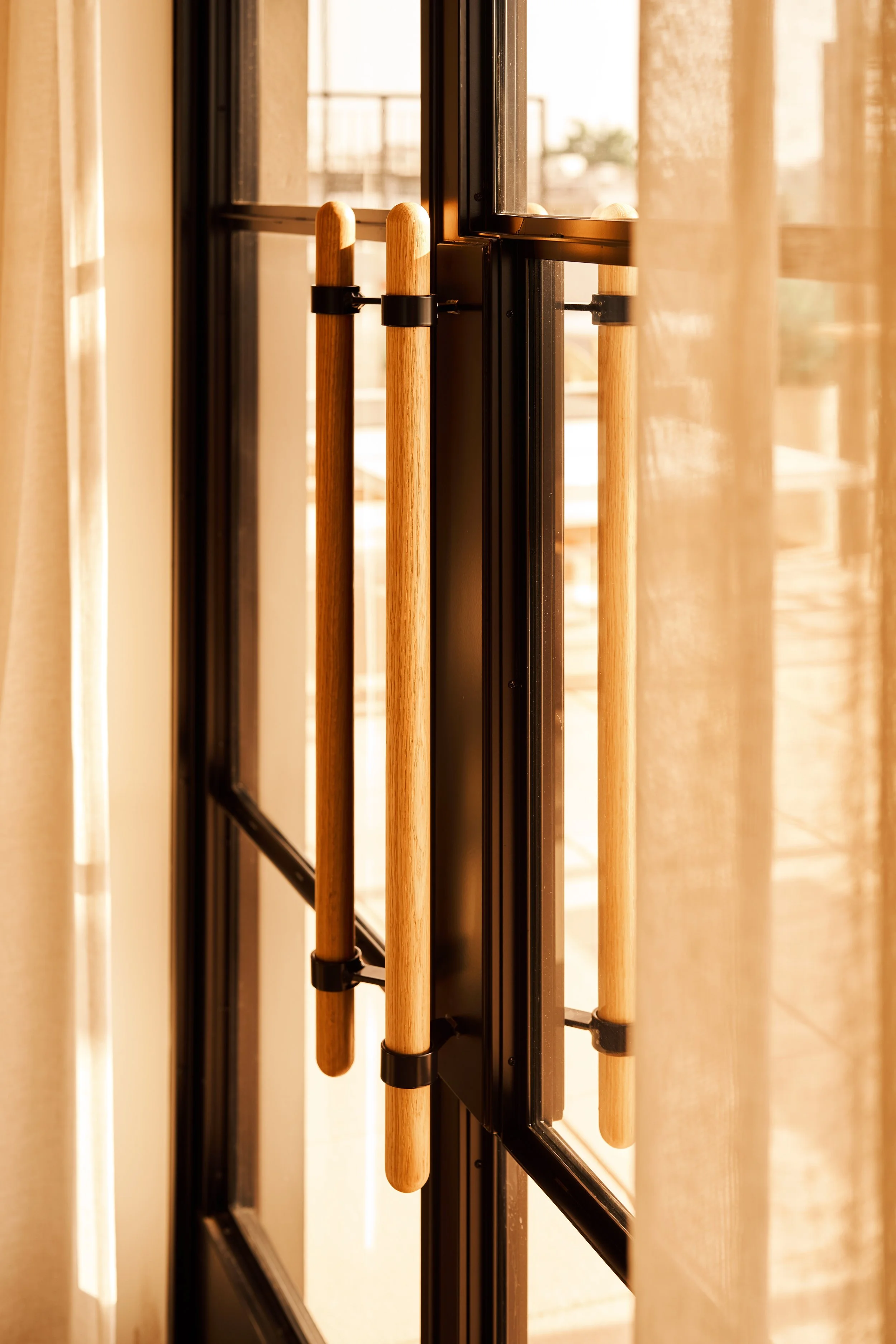 Close-up of wooden door handles on a black metal-framed glass door, with beige curtains on either side, sunlit in the background.