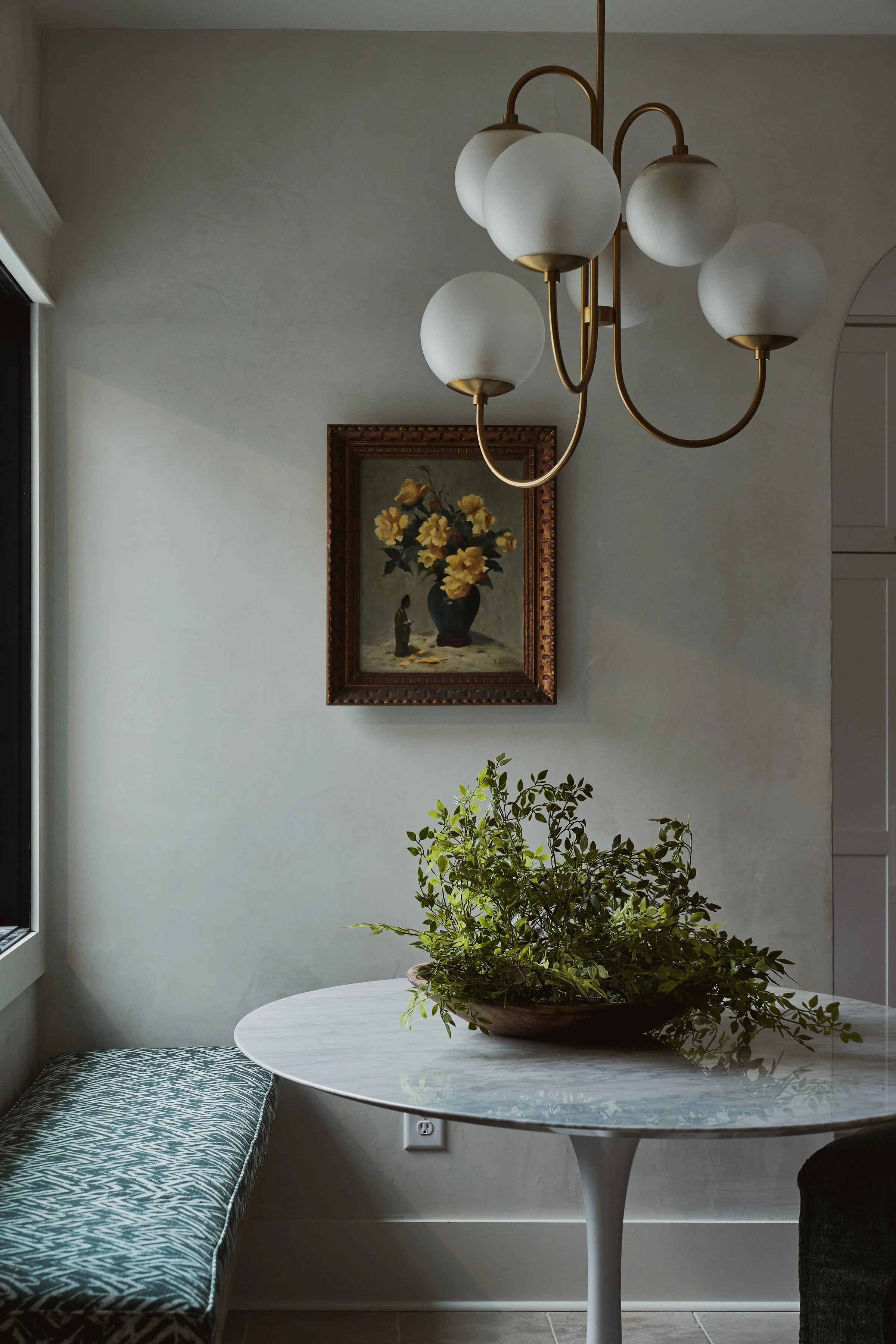 Interior room with a white round table, a green leafy plant in a brown bowl, a window, a patterned bench, a framed painting of yellow flowers in a vase, and a modern chandelier with white globes hanging from a brass frame.