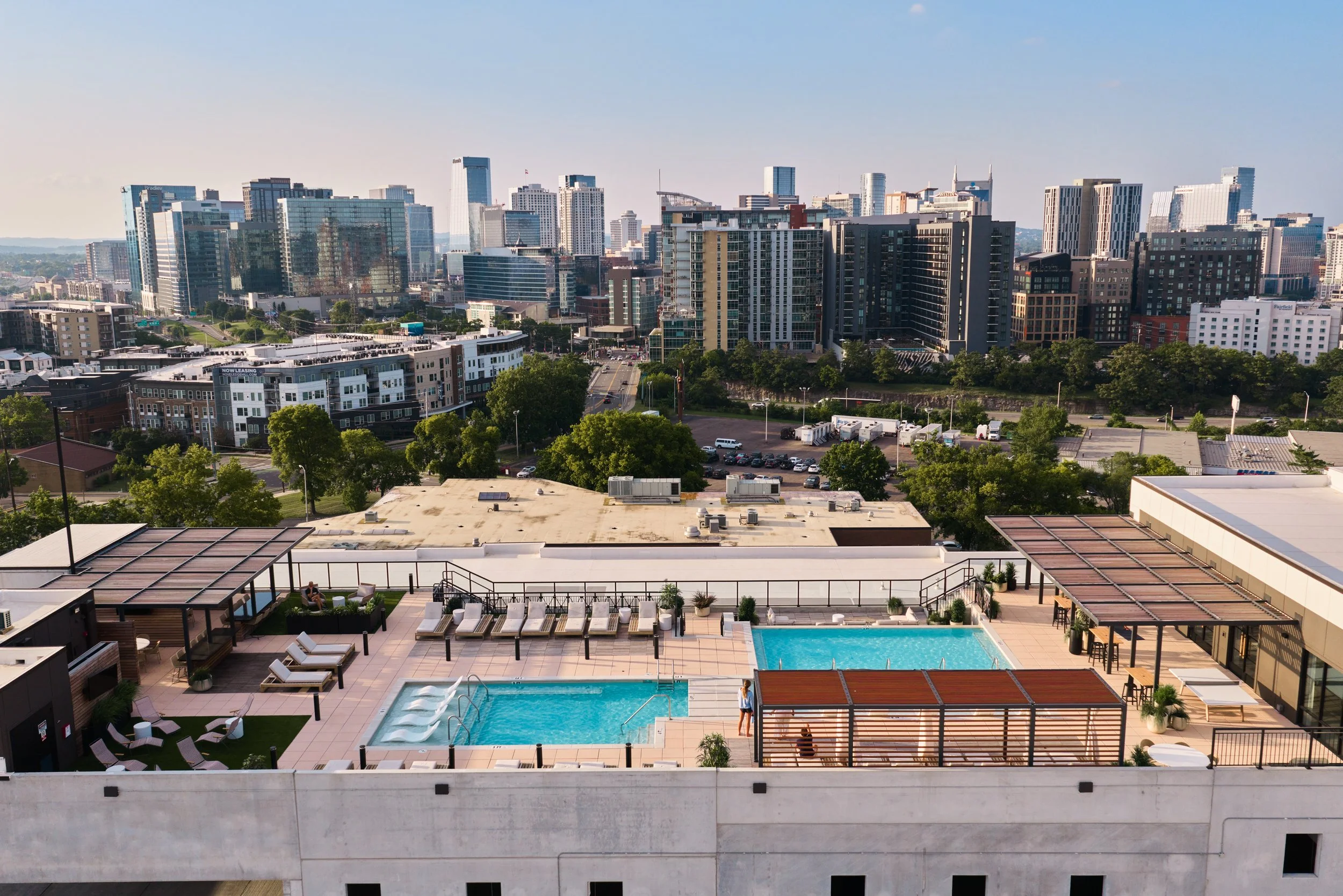 City skyline with high-rise buildings and green trees, viewed from a rooftop pool area with lounge chairs and a swimming pool in the foreground.