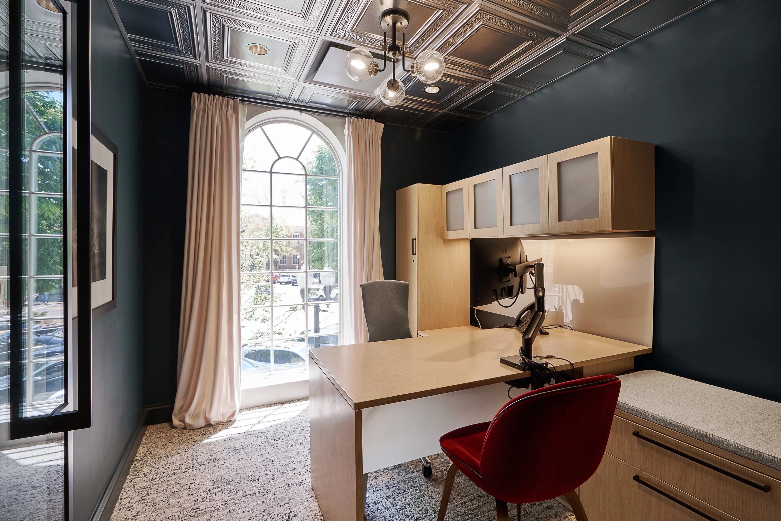 A modern office with a large arched window, dark painted walls, brushed metal ceiling tiles, and a workspace with a wooden desk, red chair, and overhead cabinets.