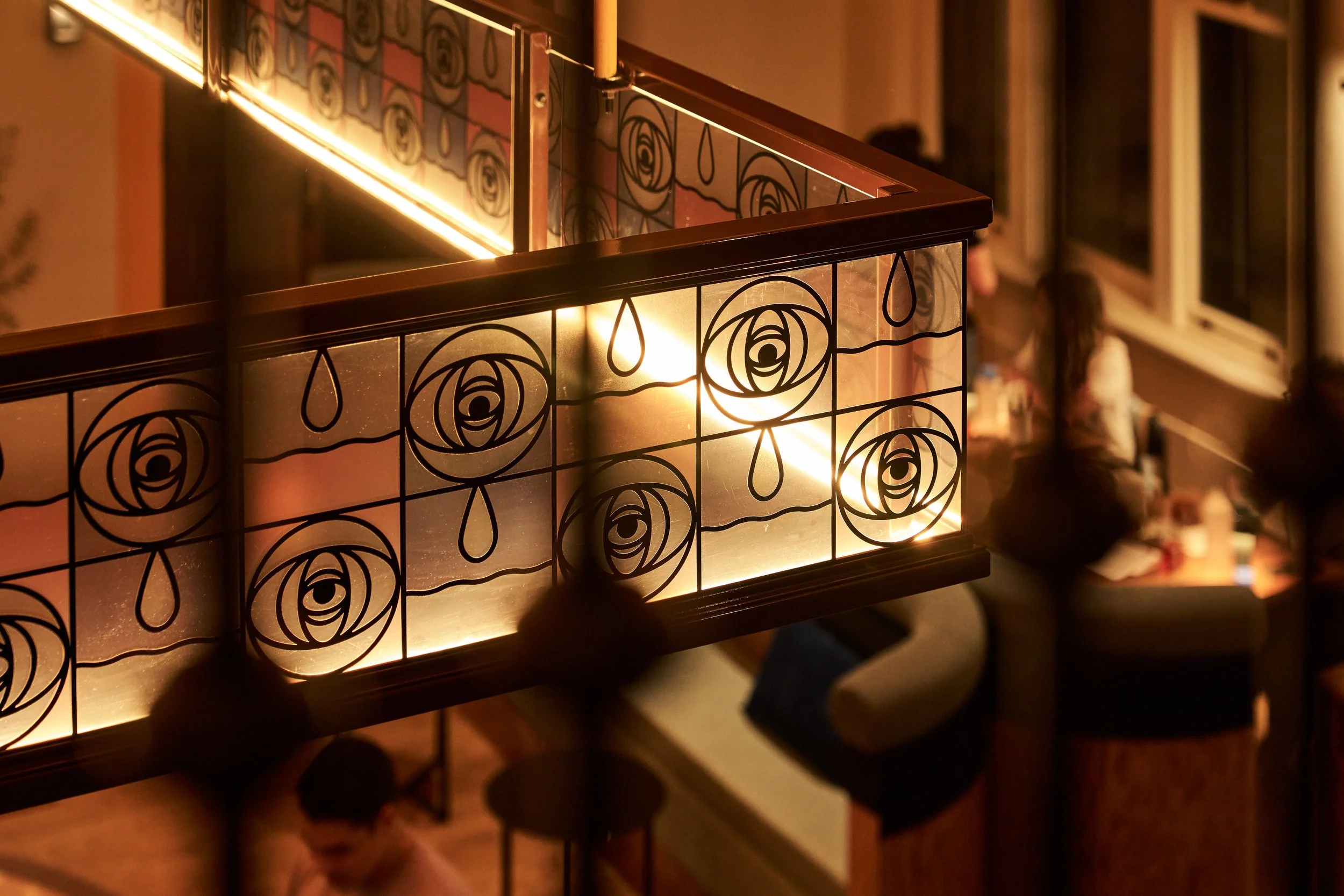 A decorative metal panel with a floral pattern illuminated from behind, creating a warm glow. The panel has a grid of roses and teardrop shapes, and is part of a railing or divider in an indoor setting, with a blurred background of people and windows