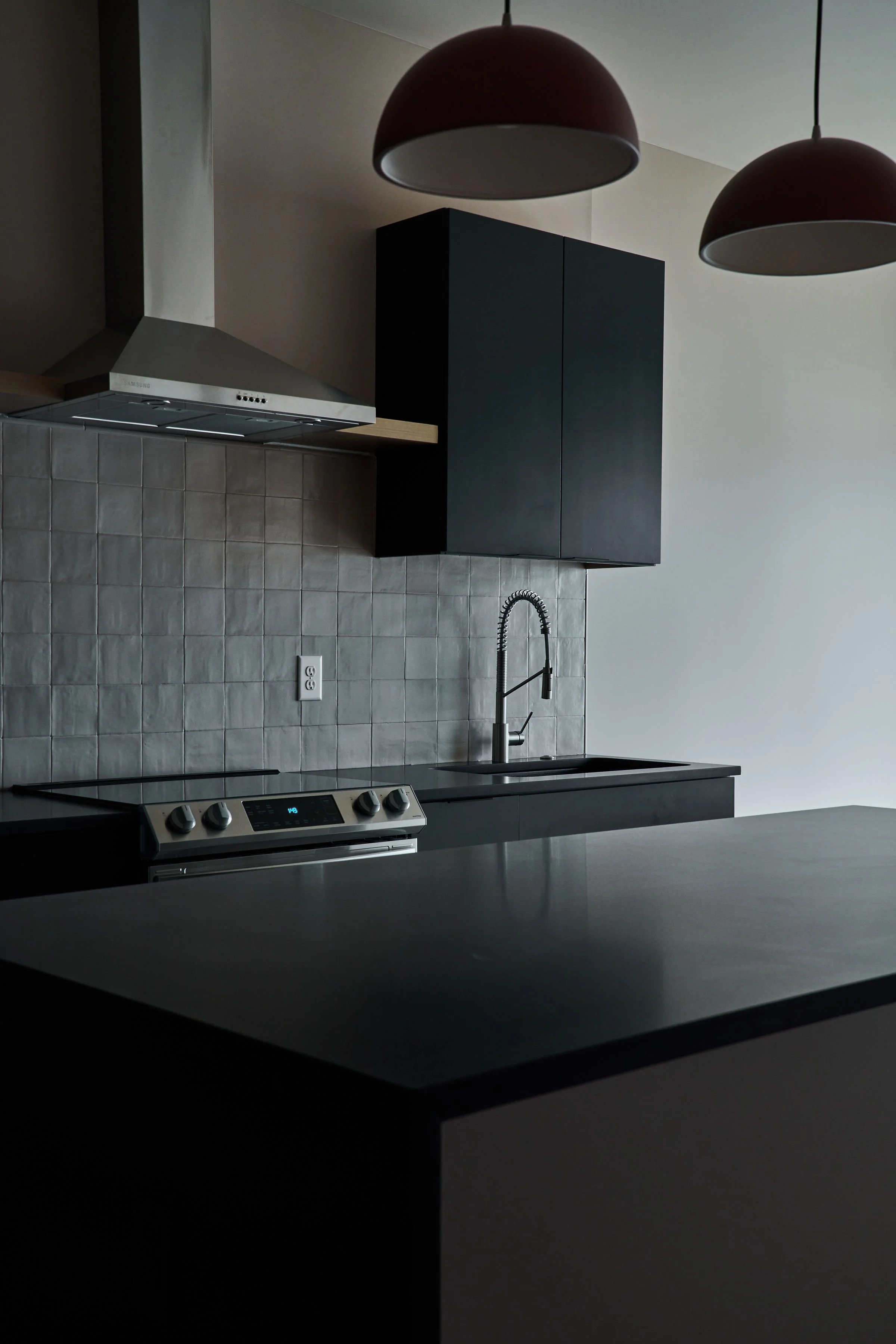 Modern kitchen with black cabinets, a white and gray tiled backsplash, a stainless steel range hood, a black stove with digital controls, and a black countertop with a sink and faucet. Two large, dark red pendant lights hang from the ceiling.