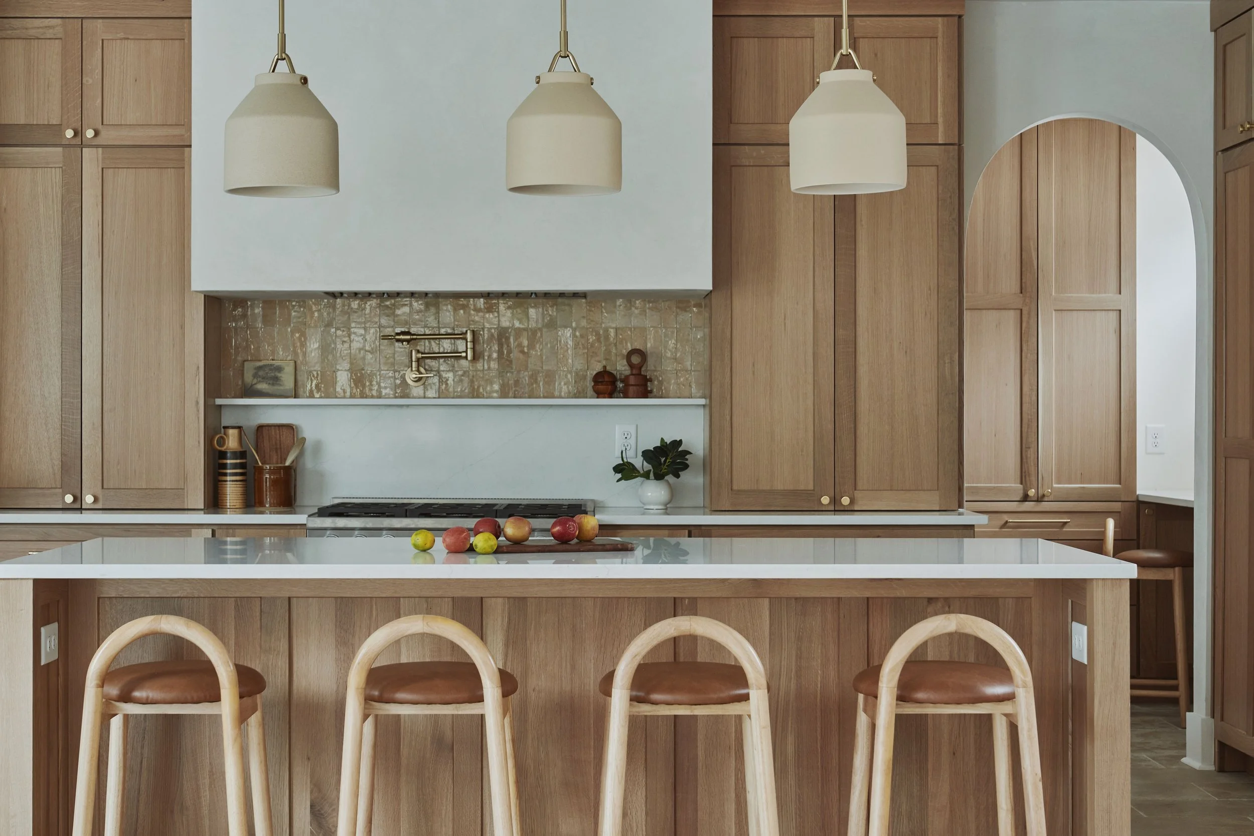Modern kitchen with wooden cabinets, a white countertop, and a table with a bowl of mixed apples. Four wooden chairs with leather seats are placed around the island. Pendant lights hang over the kitchen area.