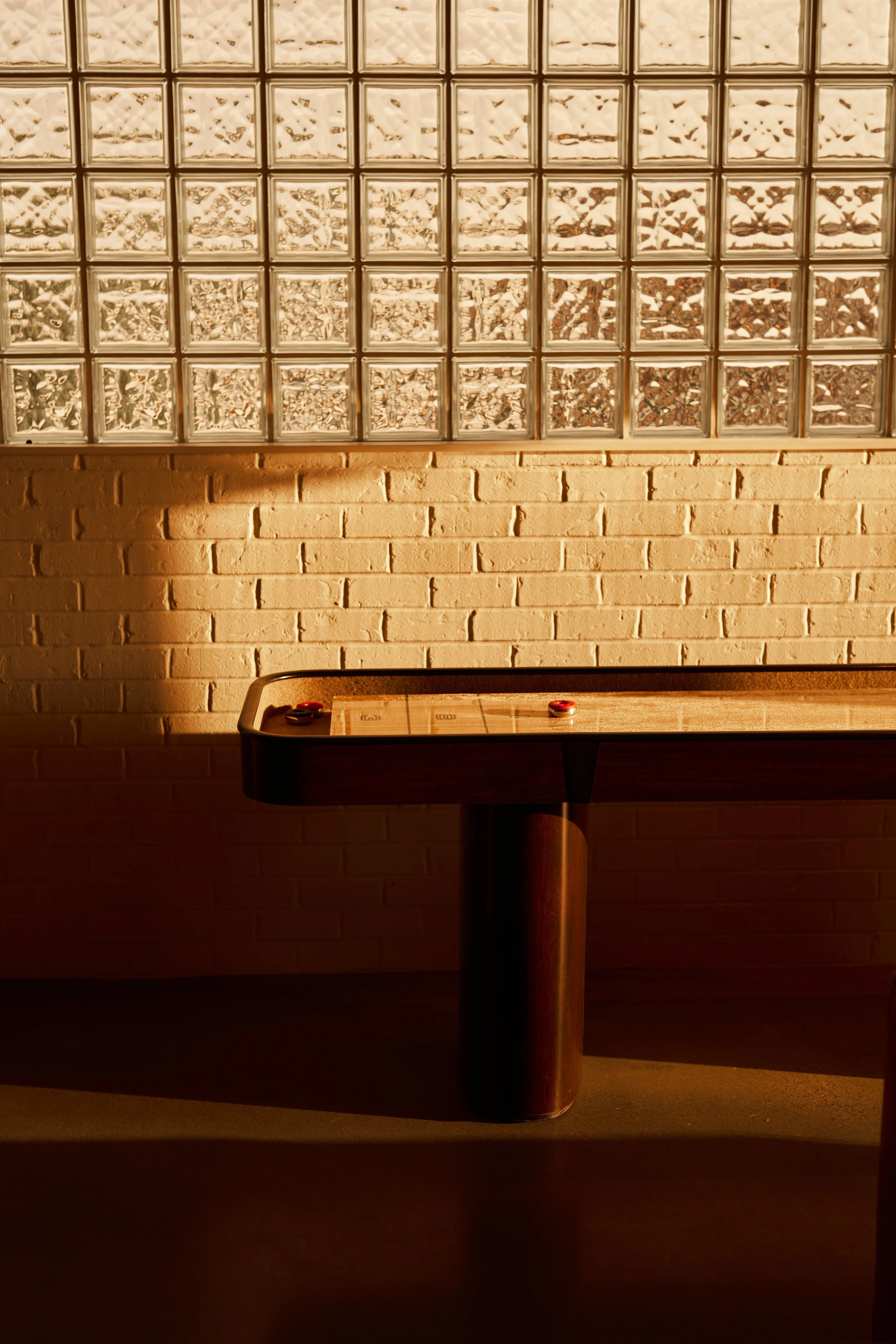 A glass block wall above a brick wall. Below the brick wall is a shuffleboard table with pucks on it.