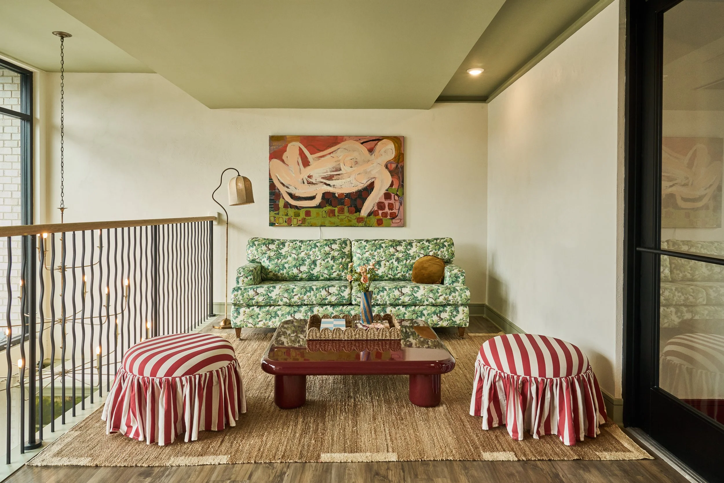 Living room with a green floral sofa, two red and white striped skirted chairs, a wooden coffee table, wall art of abstract nude figures, a small side table with flowers, and a hanging light fixture.