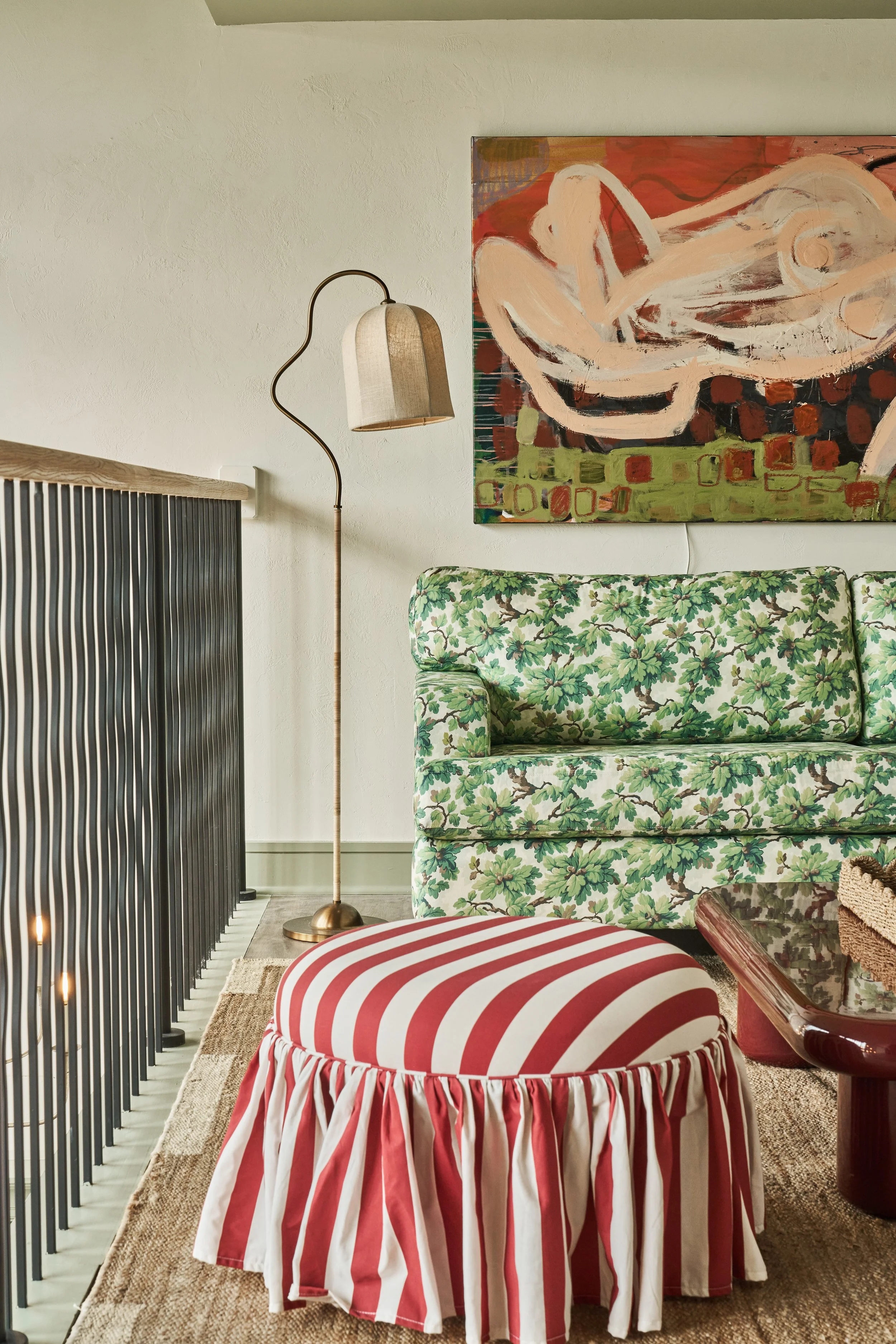 A cozy living room corner with a striped red and white ottoman, a floral-patterned green sofa, a tall standing lamp with a beige shade, a colorful abstract painting on the wall, and a slatted black room divider on the left.