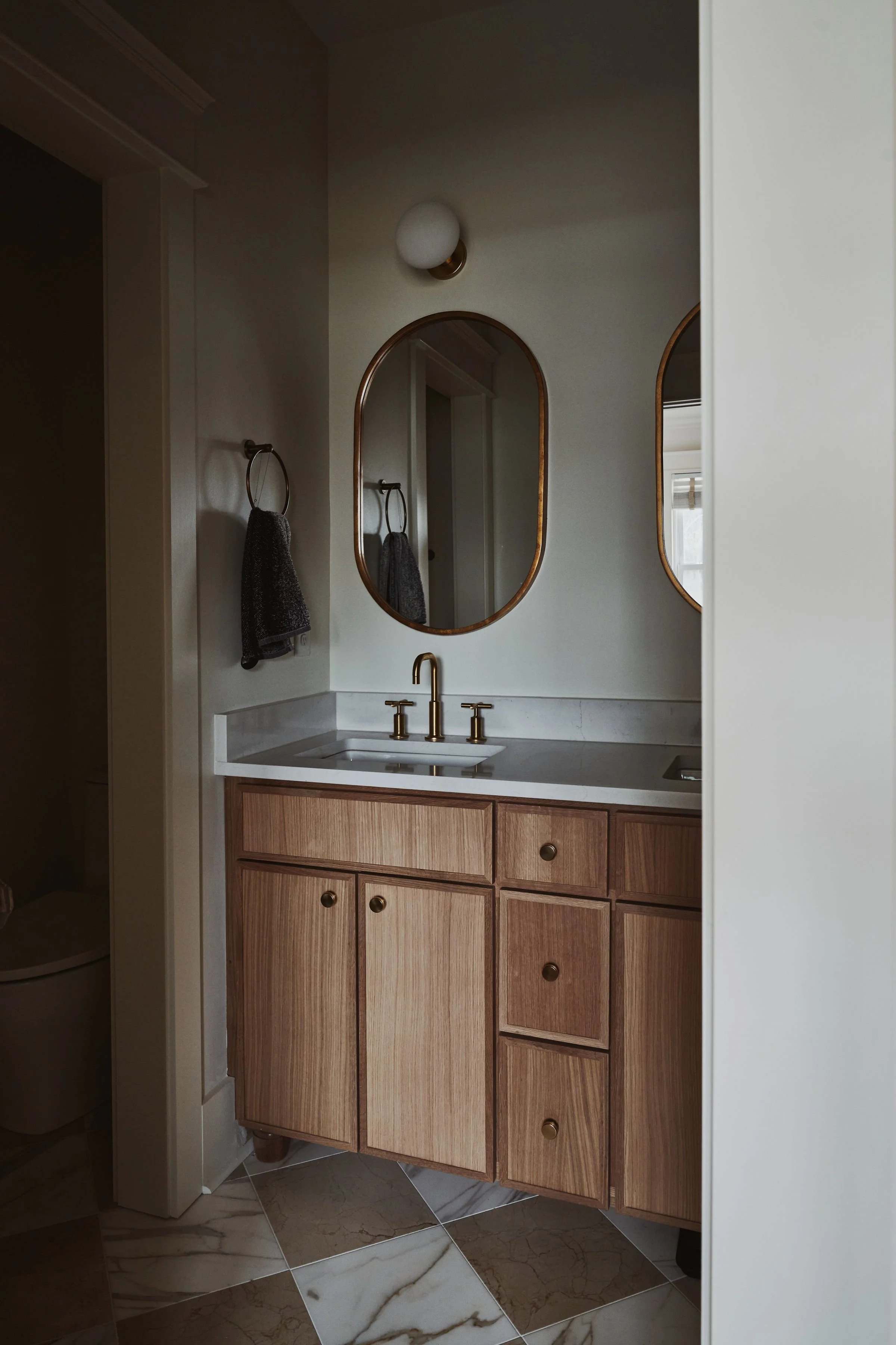 Bathroom vanity with wooden cabinets, a white marble countertop, a gold faucet, two mirrors with gold frames, a wall light above, a towel ring with a dark towel, tiled floor with marble patterns.