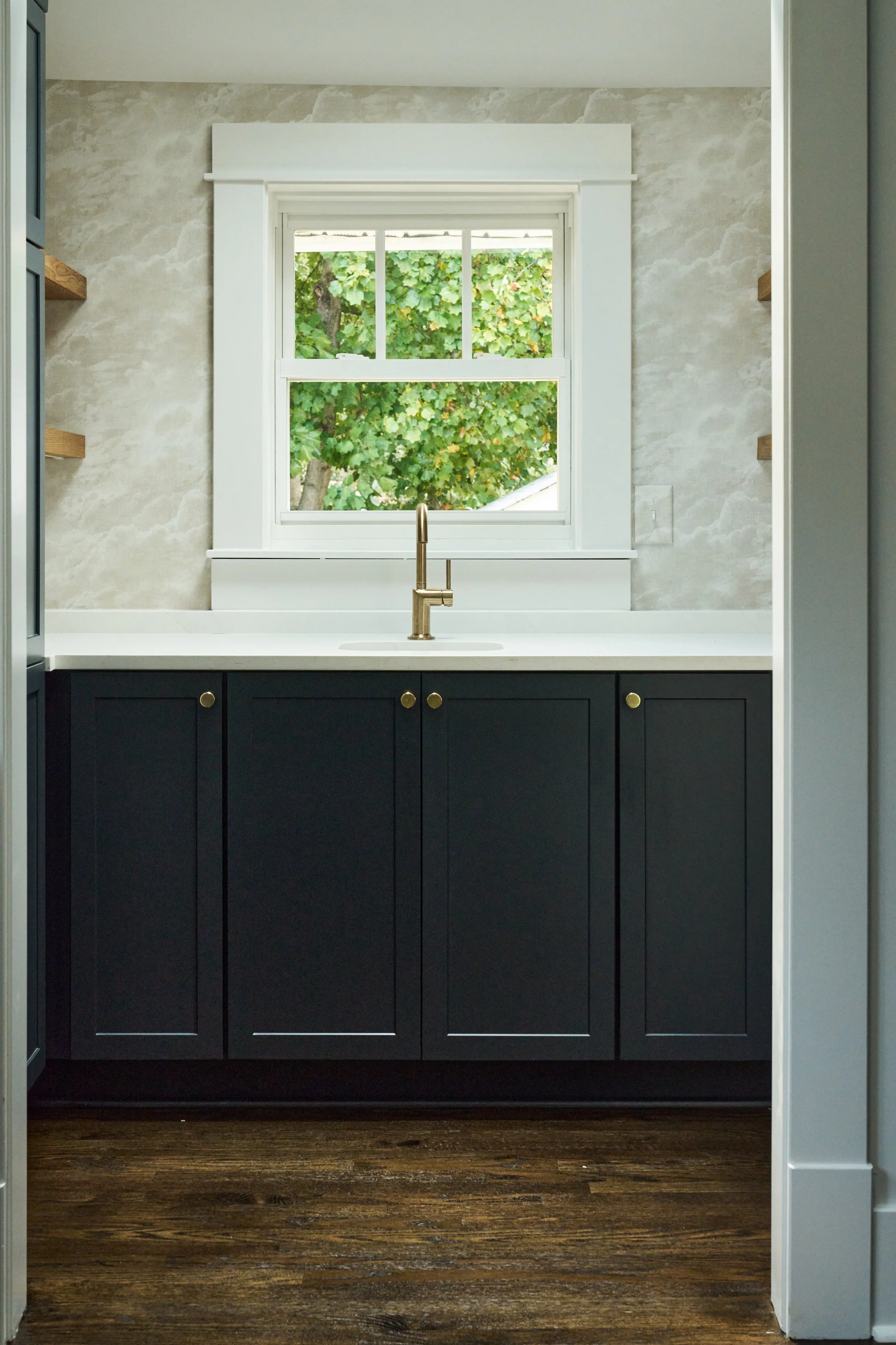 View of a kitchen window with view of green leafy tree outside, black cabinets below the window, brass sink faucet, and dark wood floor.