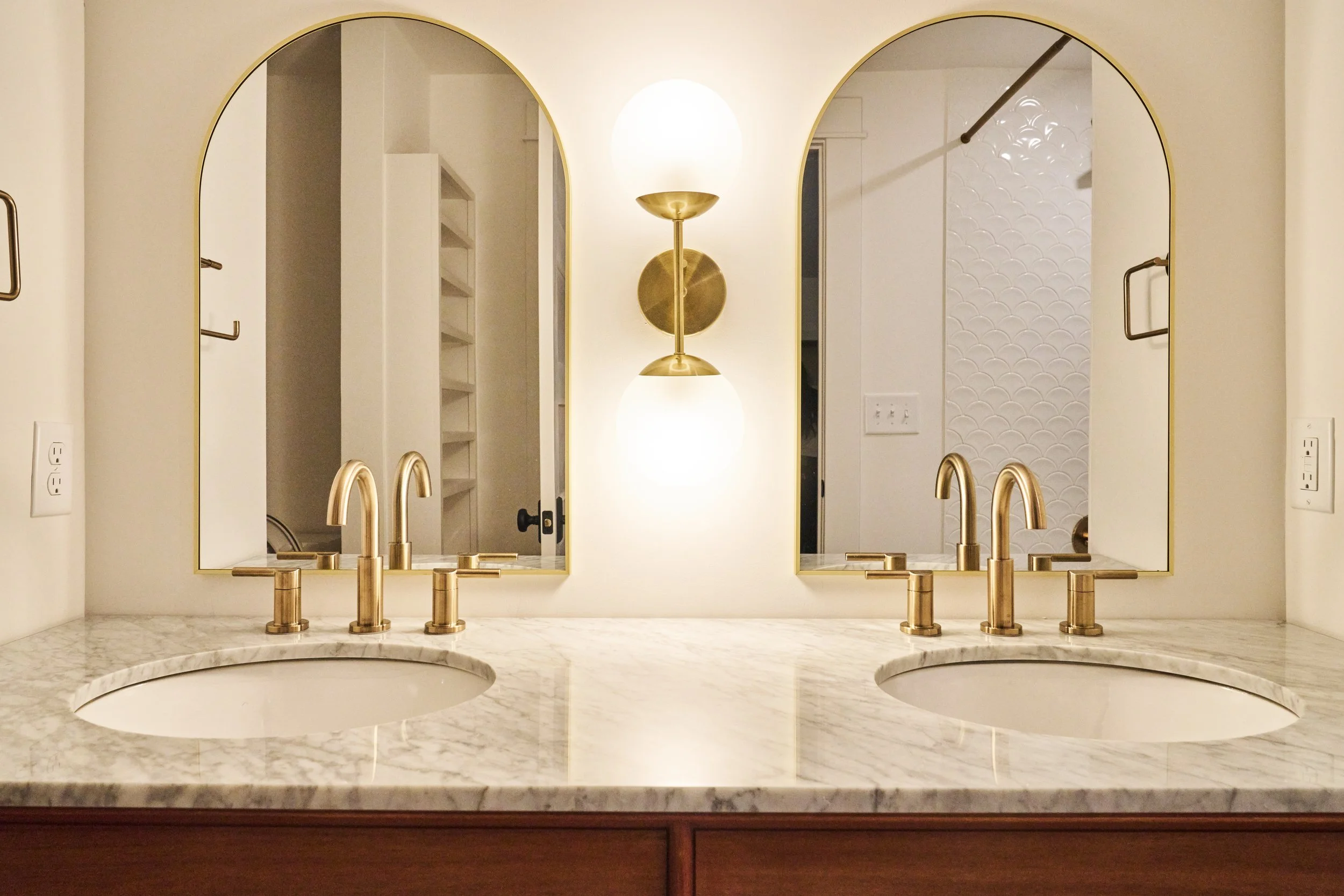 Double bathroom vanity with gold fixtures and two mirrors, marble countertop, and a wall sconce between the mirrors.