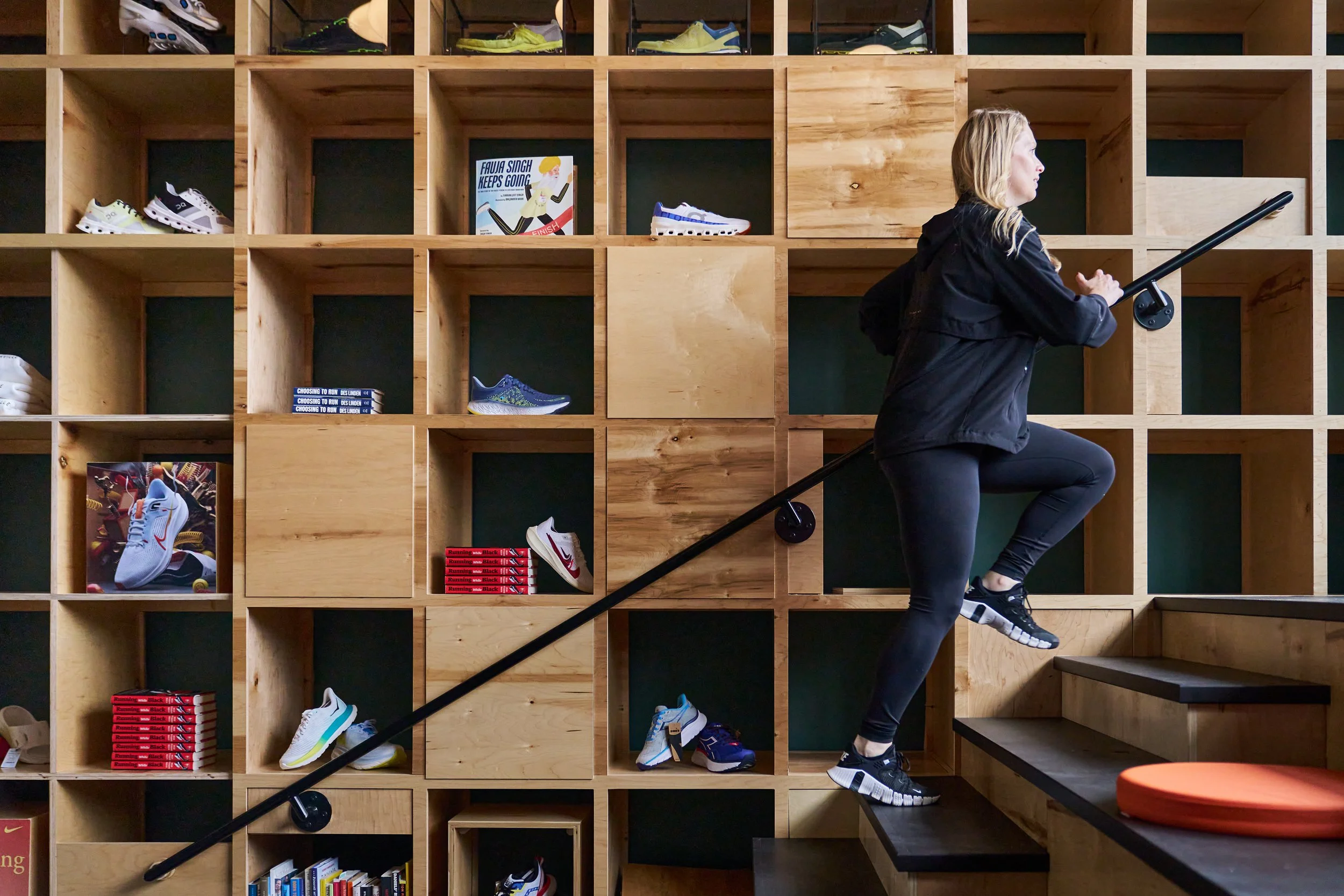 A woman in athletic clothing stepping up a stair in front of a wooden cubby wall filled with various athletic shoes and booklets.