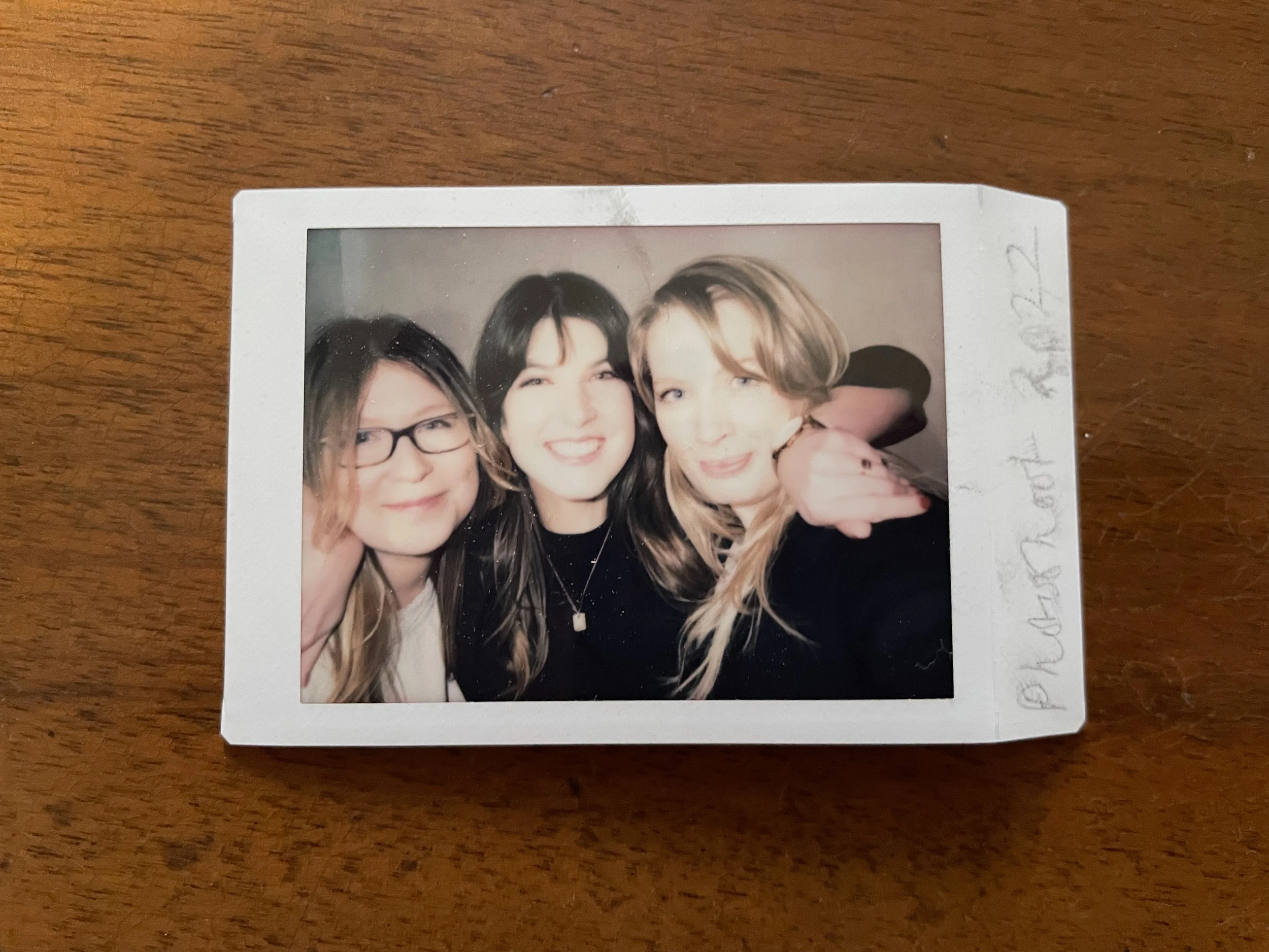 Photograph of three women smiling and posing together at a social event. The woman on the left has light brown hair and glasses, the woman in the center has dark hair, and the woman on the right has blonde hair. They are close together, with the woman on the right hugging the middle woman.