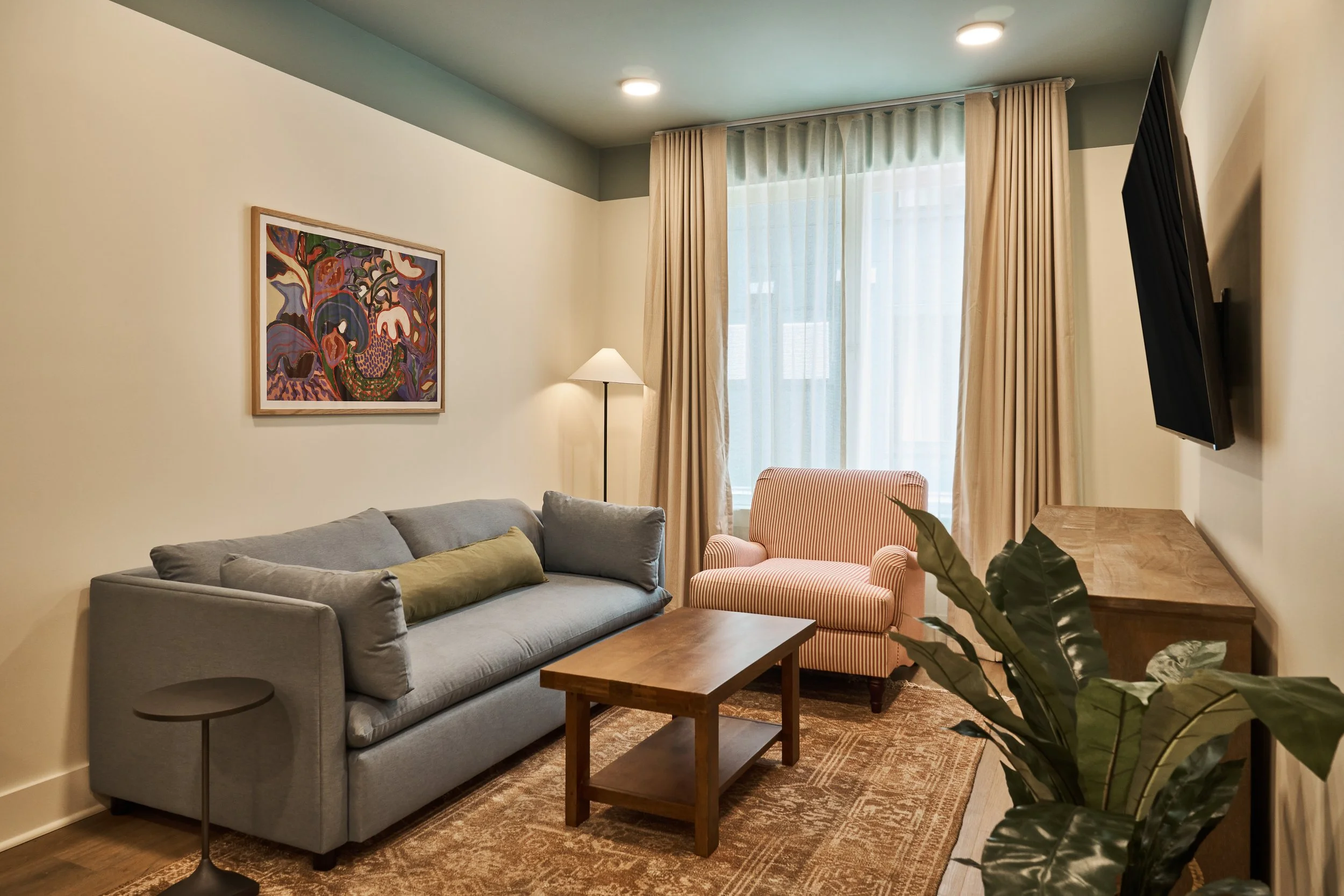 A room with a gray sofa, a pink-striped armchair, a wooden coffee table, a side table, a colorful abstract painting on the wall, a floor lamp, a large window with sheer white curtains and beige drapes, and a television mounted on the wall.