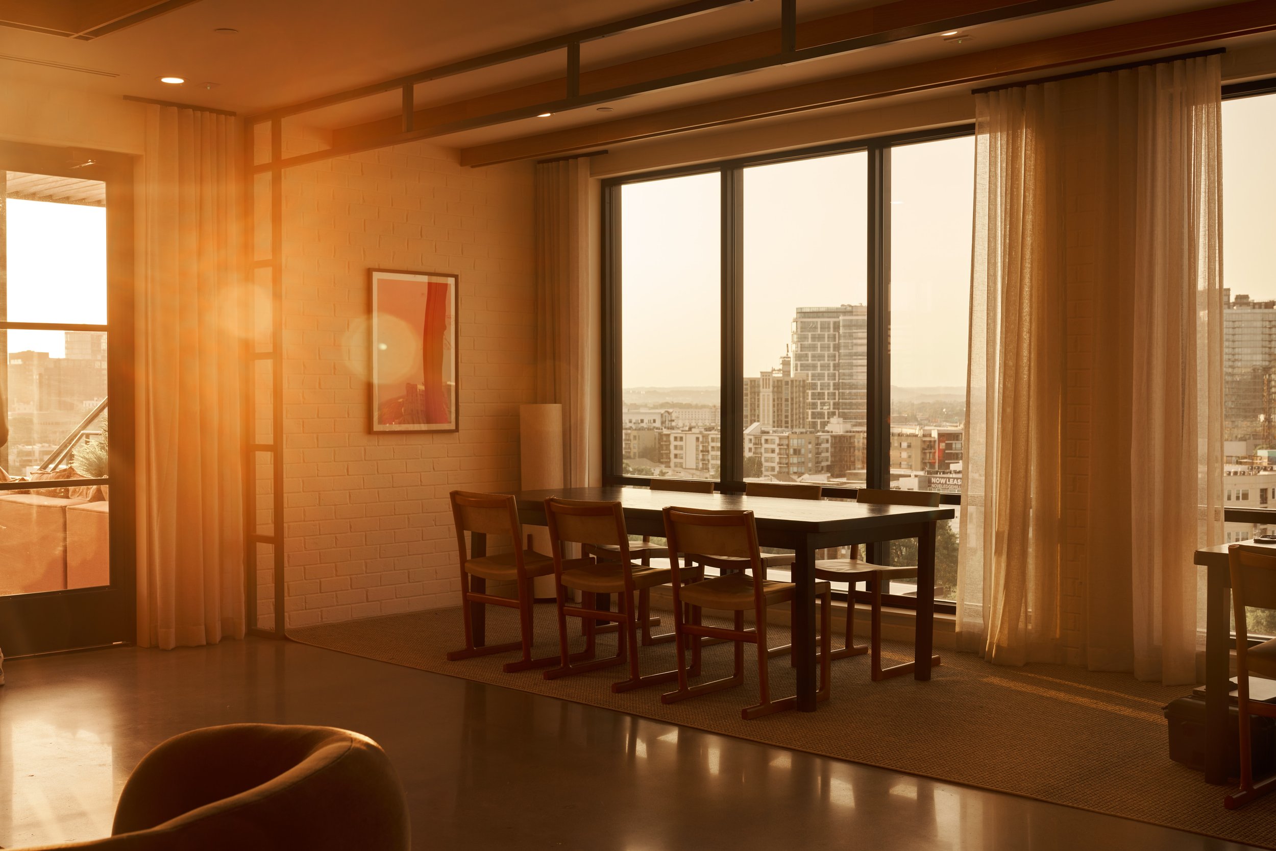 Sunlight streams through large windows in a modern apartment, illuminating a dining table with chairs and a piece of abstract artwork on a brick wall.