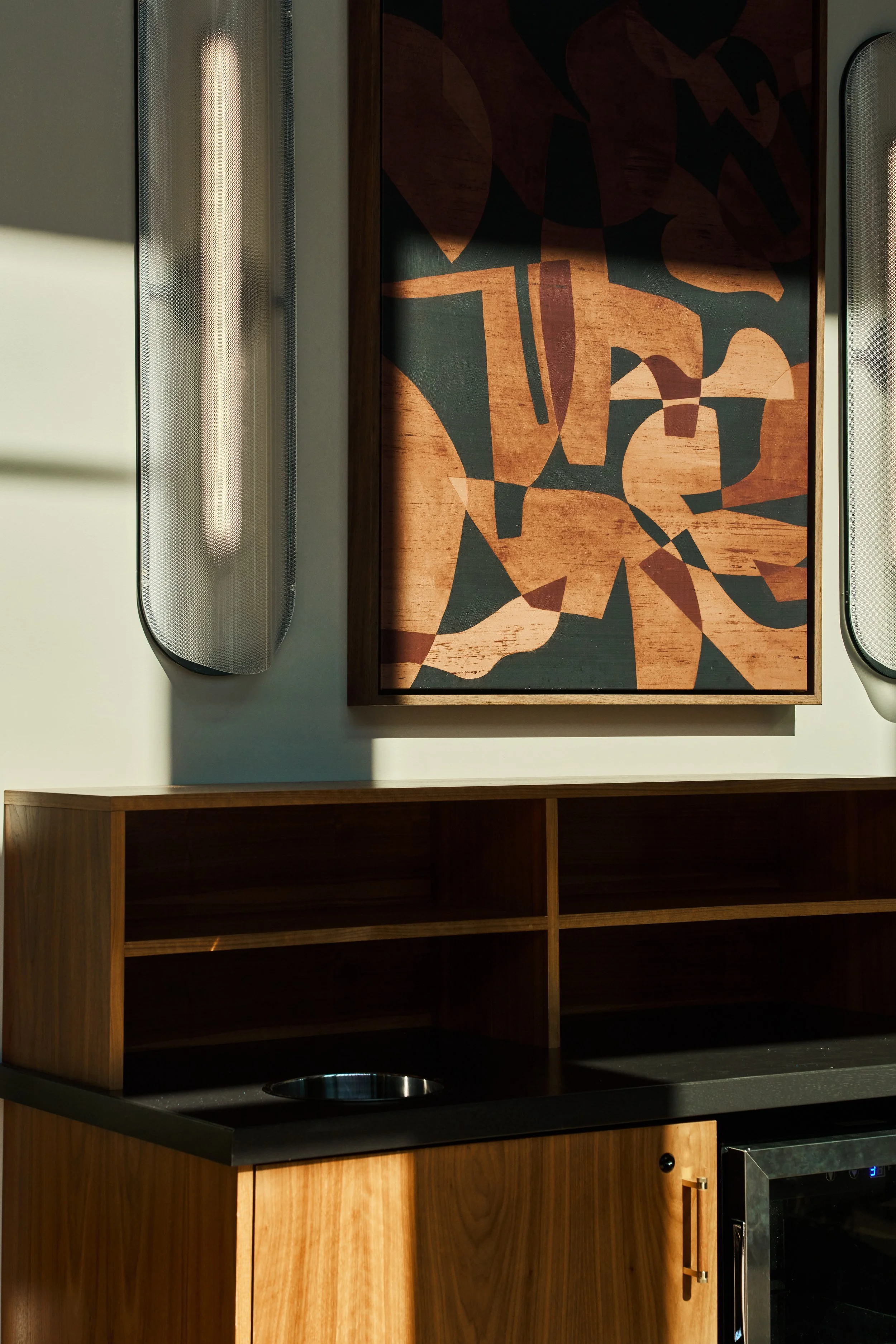 A modern kitchen cabinet with open shelving, a black countertop with a small circular sink, a wooden drawer front, a wall with a large abstract art piece, and two vertical frosted glass panels on either side of the art.
