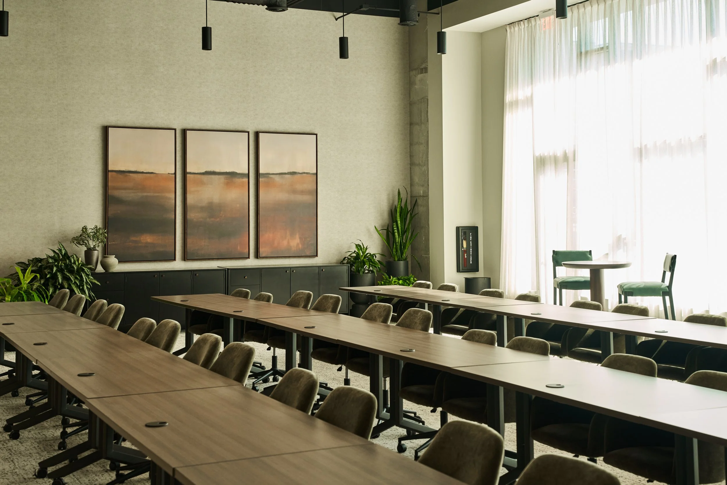 A modern conference room with long wooden tables and multiple chairs, large windows with sheer curtains, wall art, plants, and pendant lighting.