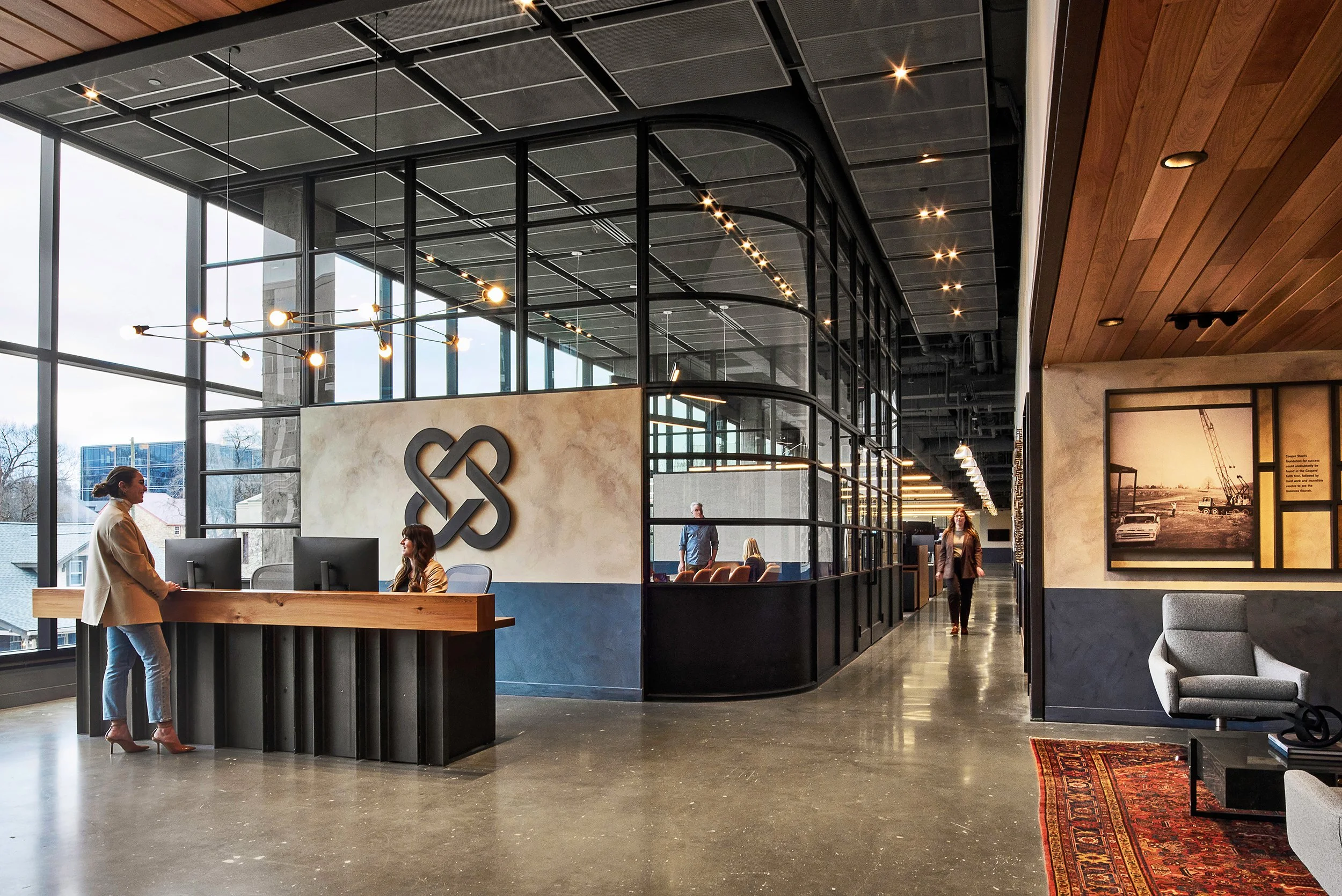 Modern office reception area with a reception desk, people talking, large glass windows, wooden ceiling, and wall art.