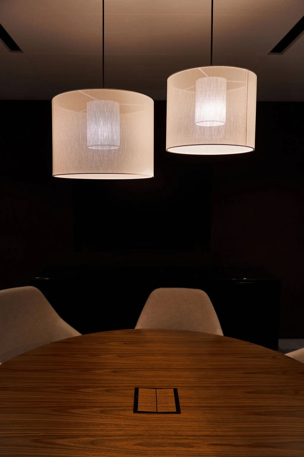 Two white, cylindrical pendant lamps hanging above a wooden conference table with beige chairs in a dimly lit room.