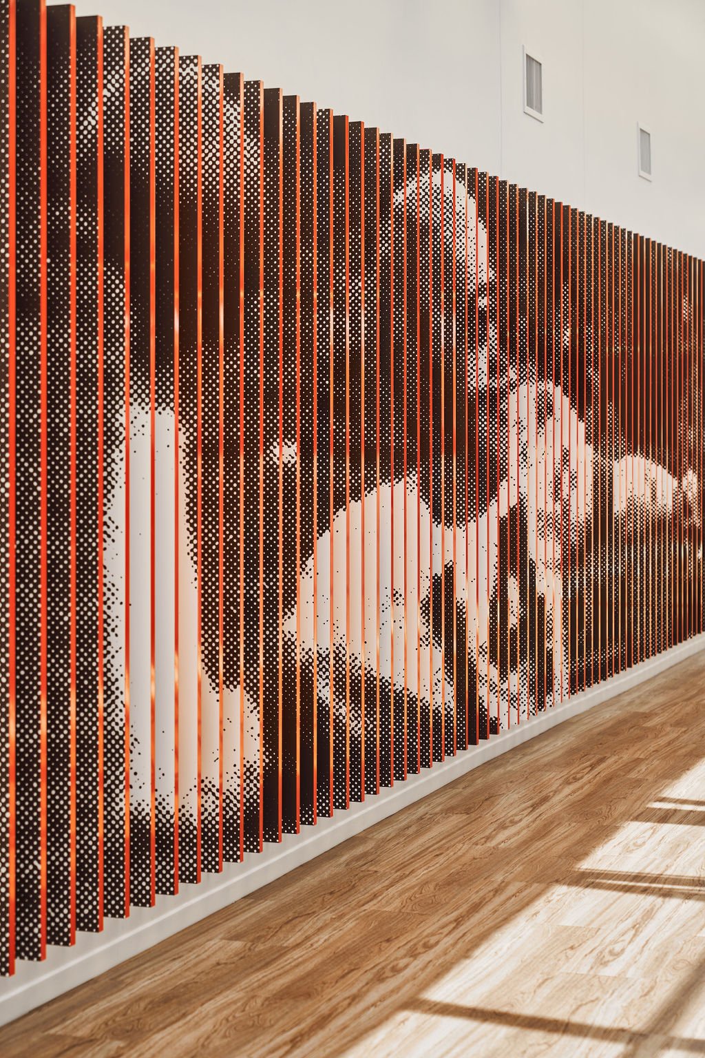 An abstract art installation featuring a large black and white face image created with small dots on a wall with black and orange vertical slats, with sunlight and shadows on a wooden floor.