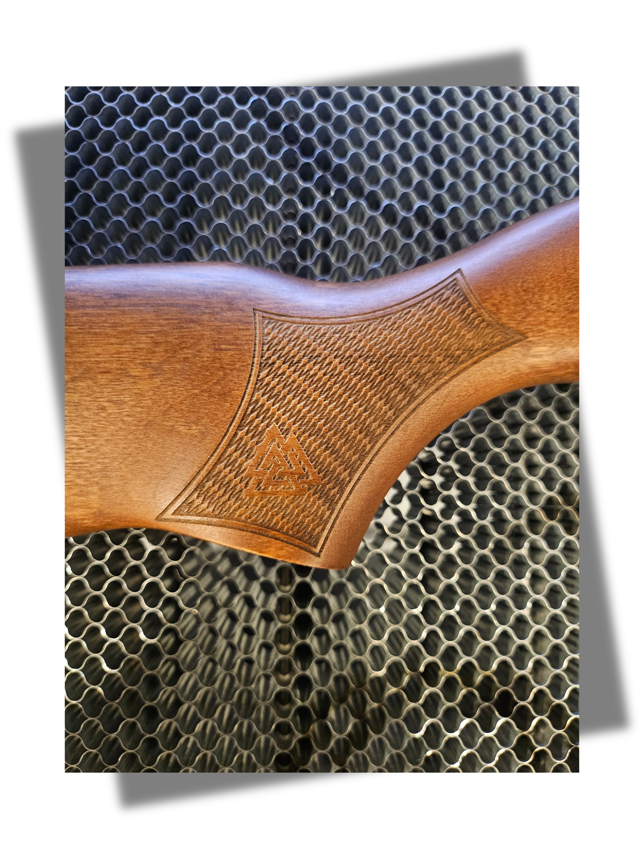 Close-up of a wooden rifle stock with an etched viking geometric pattern and logo, resting on a metal mesh surface.