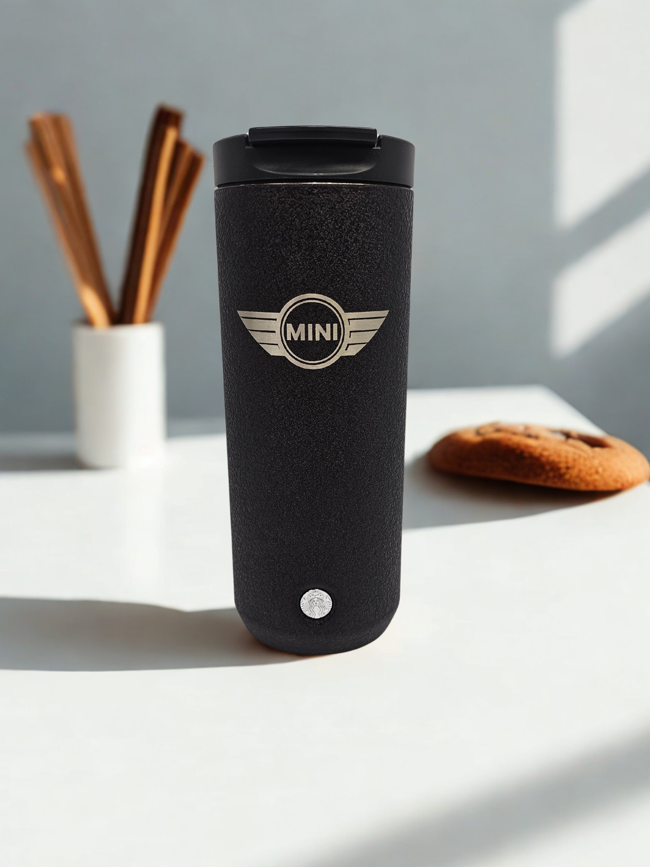 A black MINI branded travel mug on a white surface with a container of cinnamon sticks and a cookie in the background.