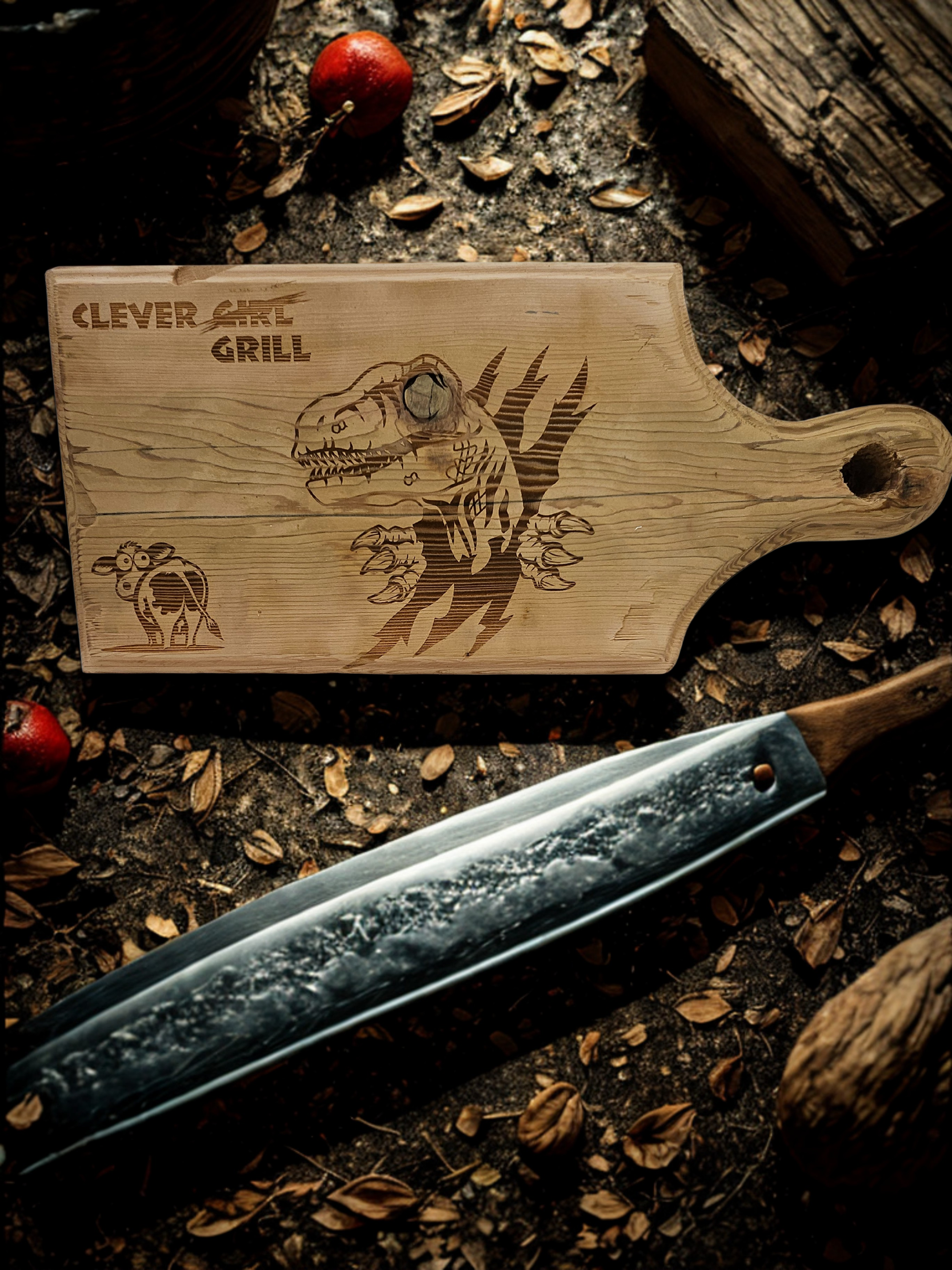 A wooden cutting board with an engraving of a dinosaur, the words 'Clever Grill,' and a small cow illustration. Next to the cutting board is a large, weathered knife on the ground, surrounded by dry leaves and a rusty log.