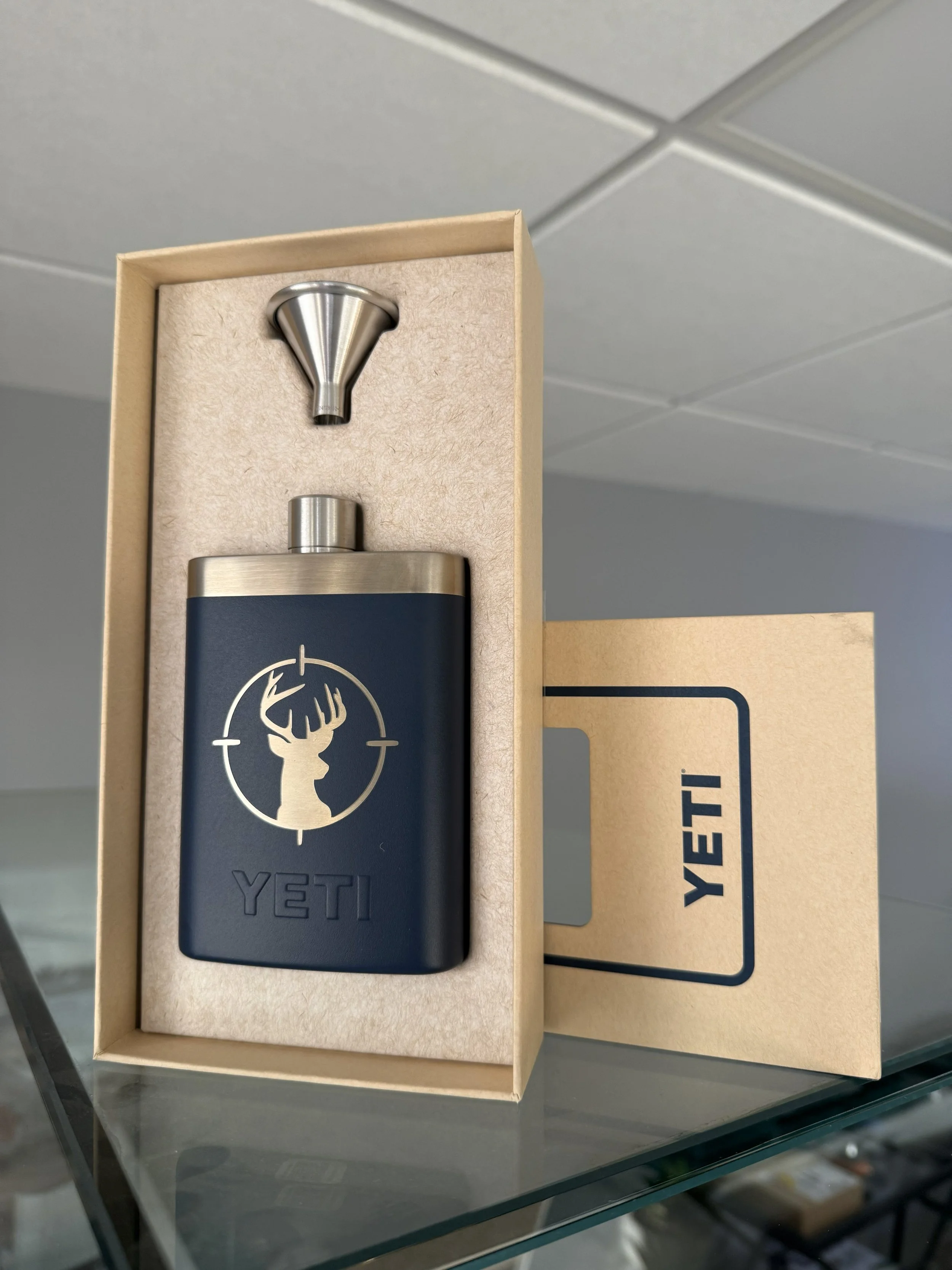 A YETI flask with a deer logo and crosshair design, in its packaging, along with a metal funnel, on a glass surface.