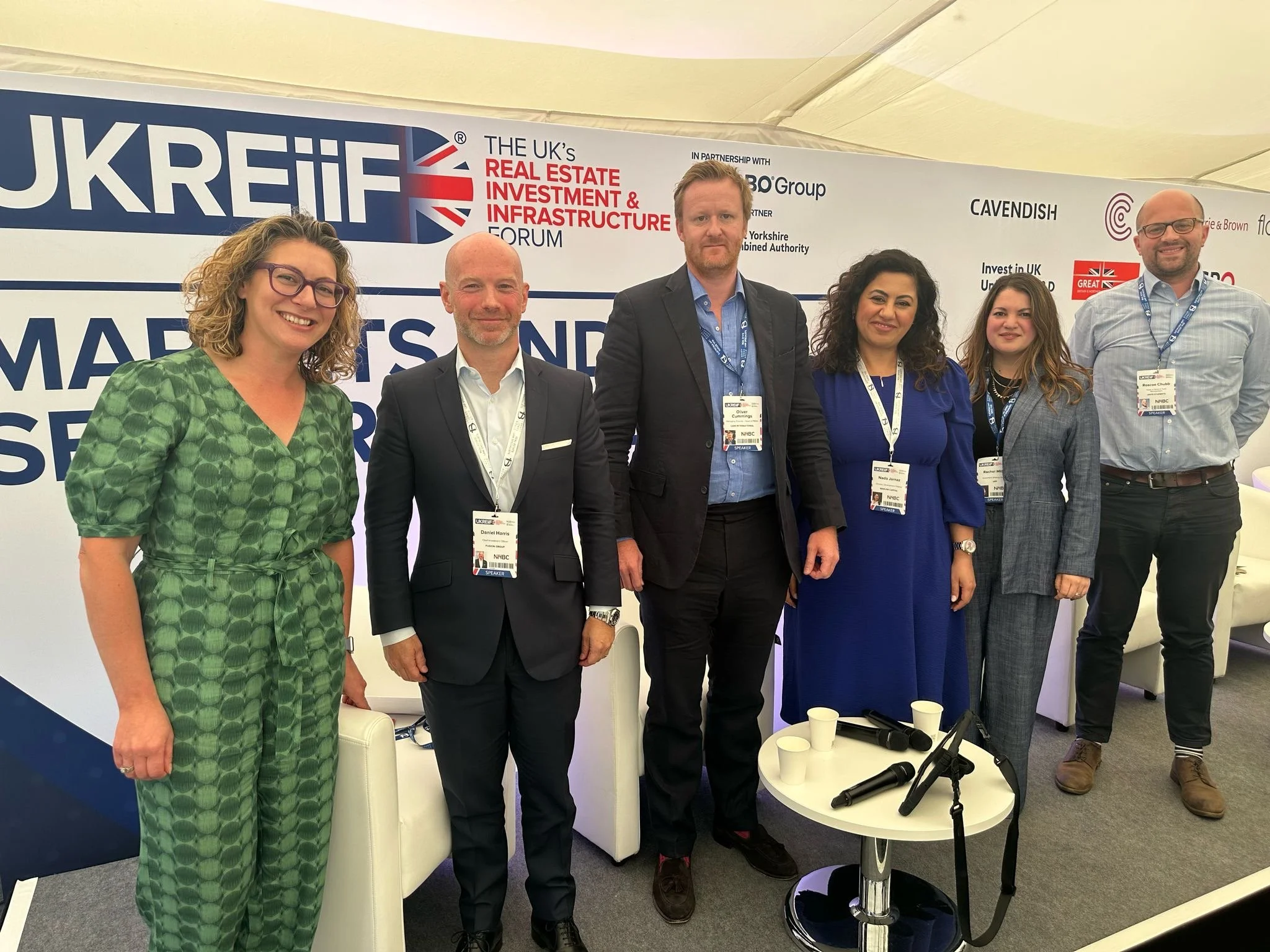 Group of six professionally dressed individuals standing side by side at a real estate forum, with a banner displaying 'UKREIF' and related logos in the background.