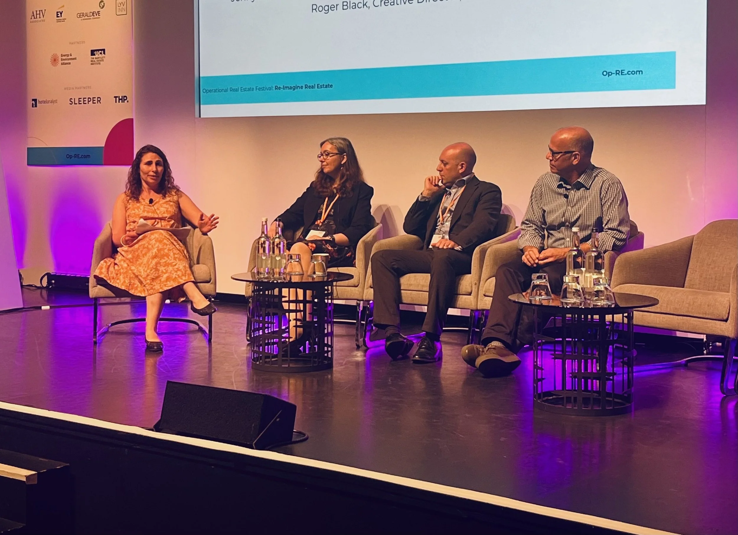 Four individuals seated on stage participating in a panel discussion at a conference, with a woman in an orange dress speaking while three others listen. There are tables with water bottles and glasses, and a large screen behind them displaying information.
