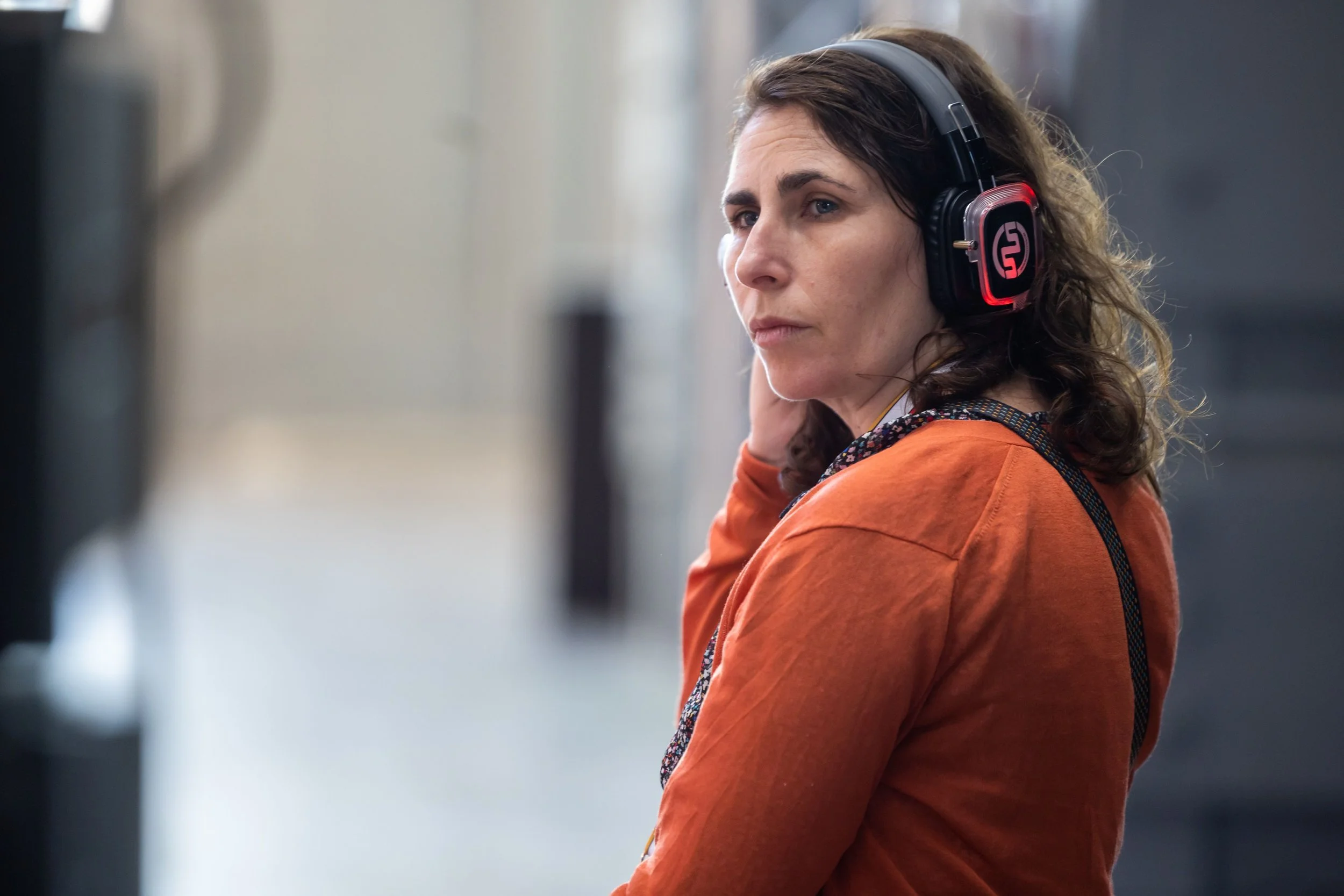 A woman with brown curly hair wearing headphones and an orange jacket, standing outdoors with a blurred background.