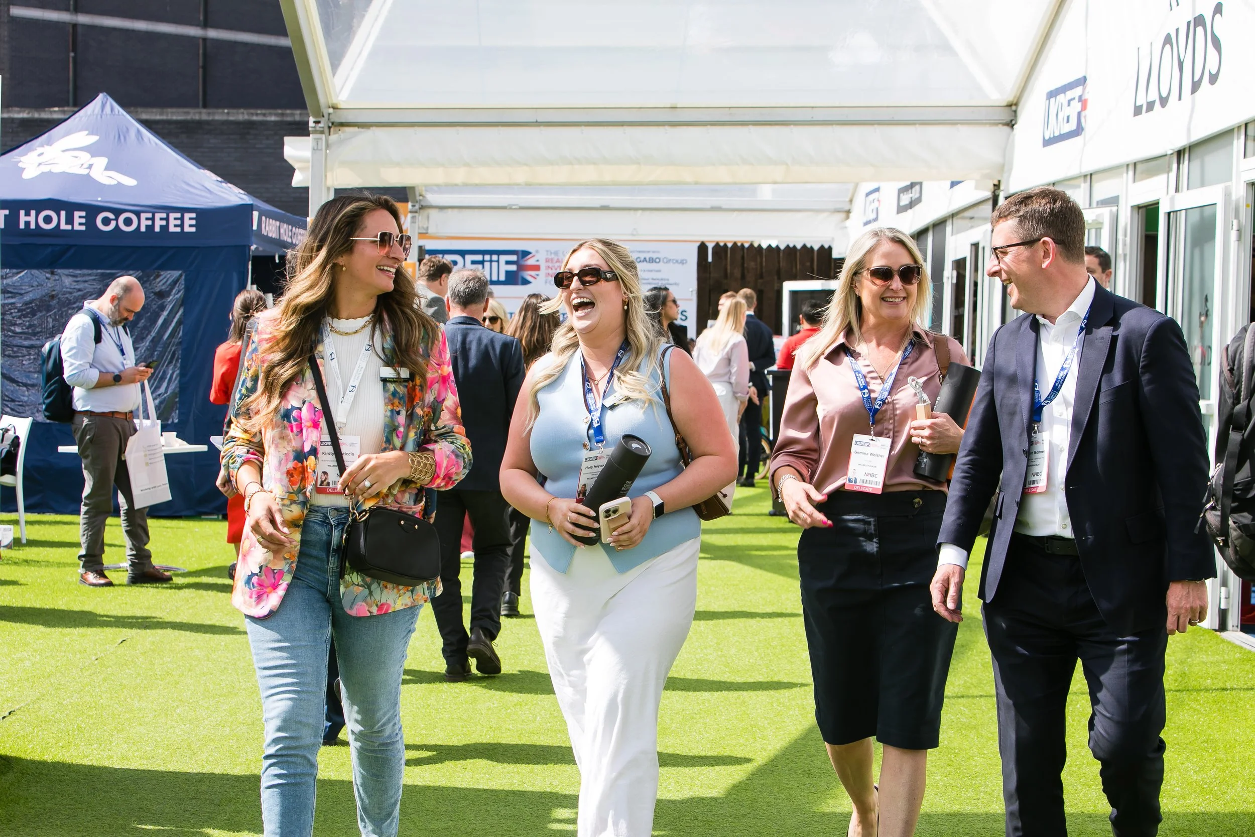 Four people walking and talking together outdoors at a professional event, with a tent and other attendees in the background.