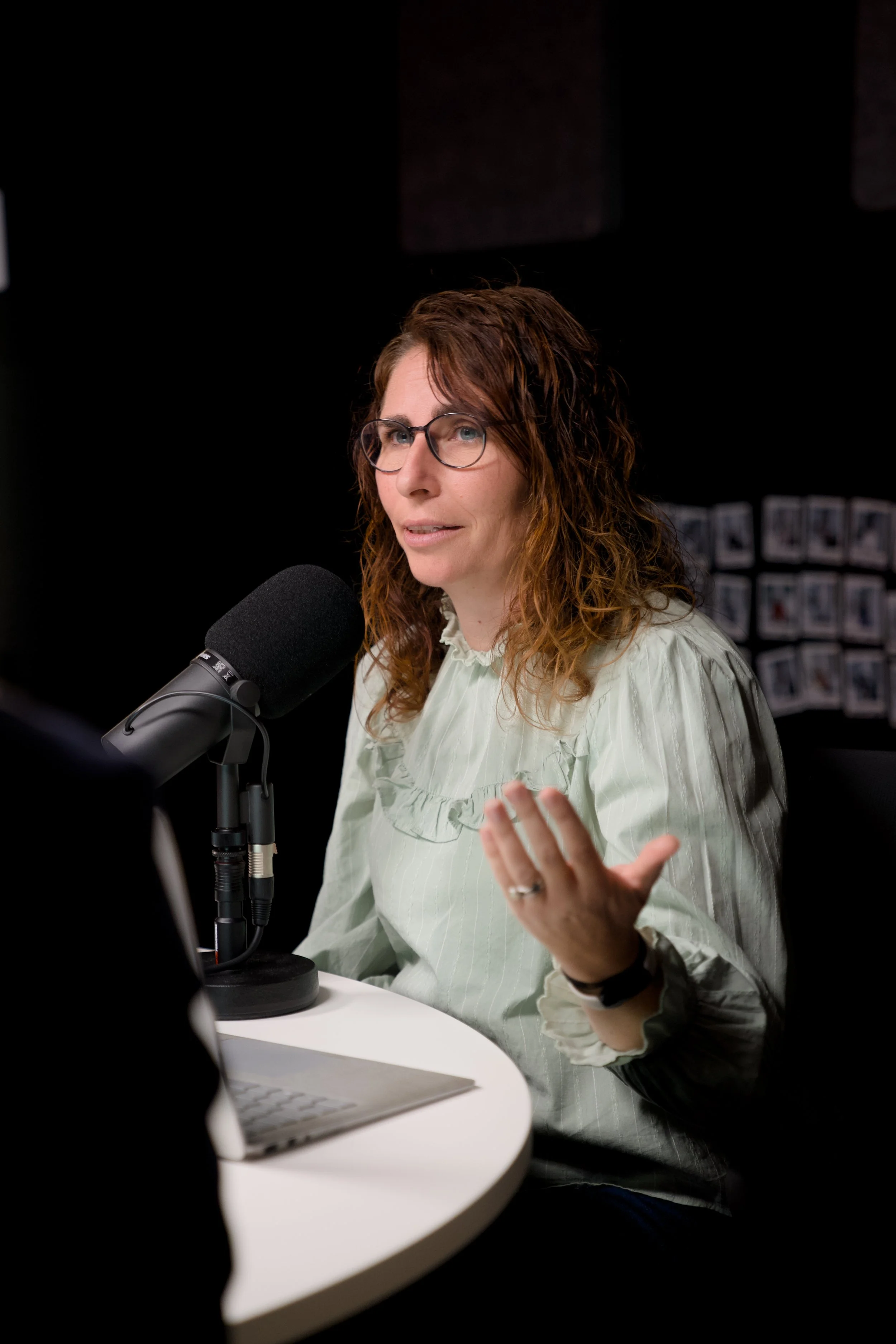 A woman with curly red hair, glasses, and a light green blouse speaking into a microphone at a podcast or radio studio.