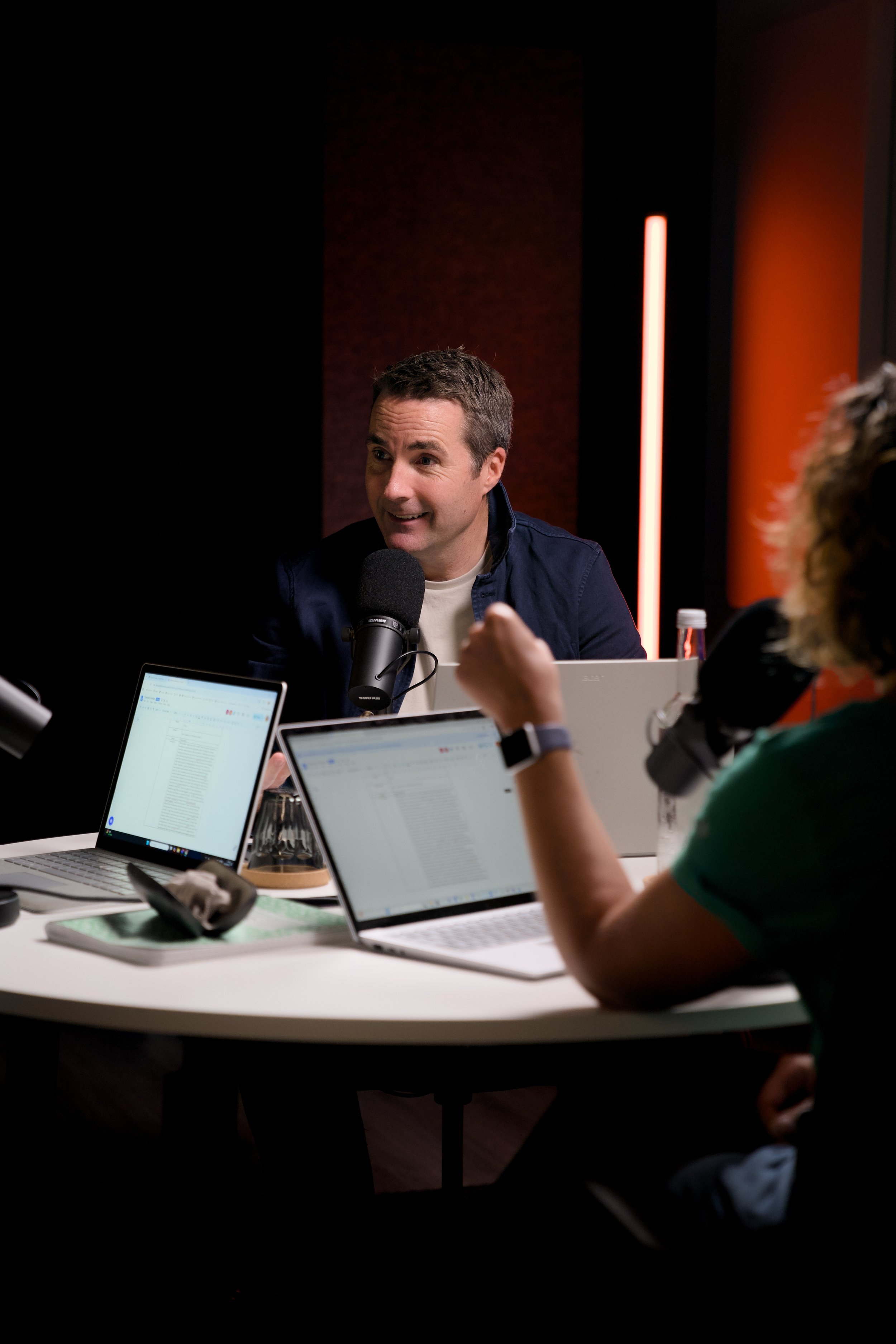 A podcast host with short dark hair speaking into a microphone during a recording session with two guests using laptops in a dimly lit studio.