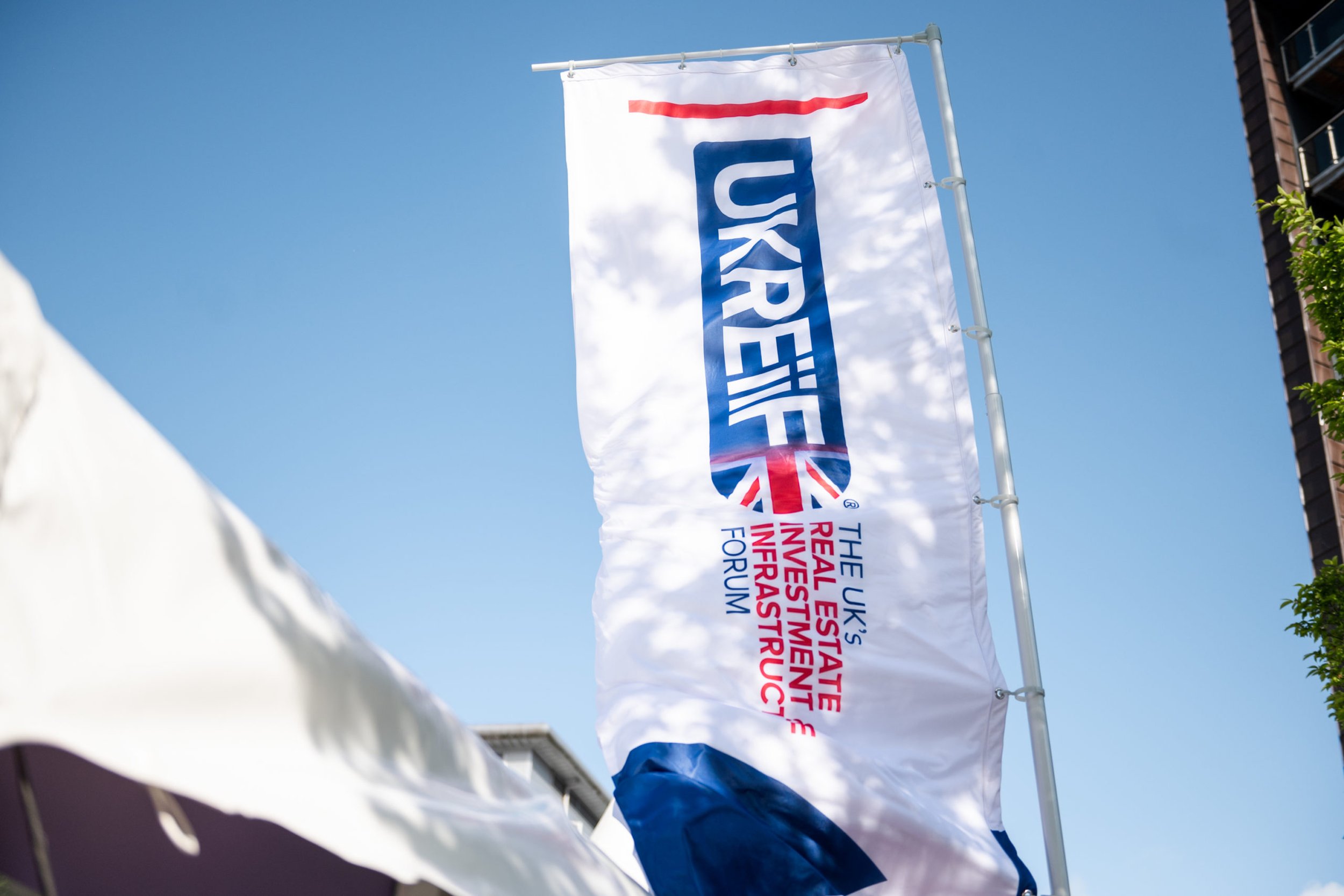 Flag with the UKREXPO logo and text 'The UK's Real Estate Investment & Infrastructure Forum' against a blue sky with some clouds.