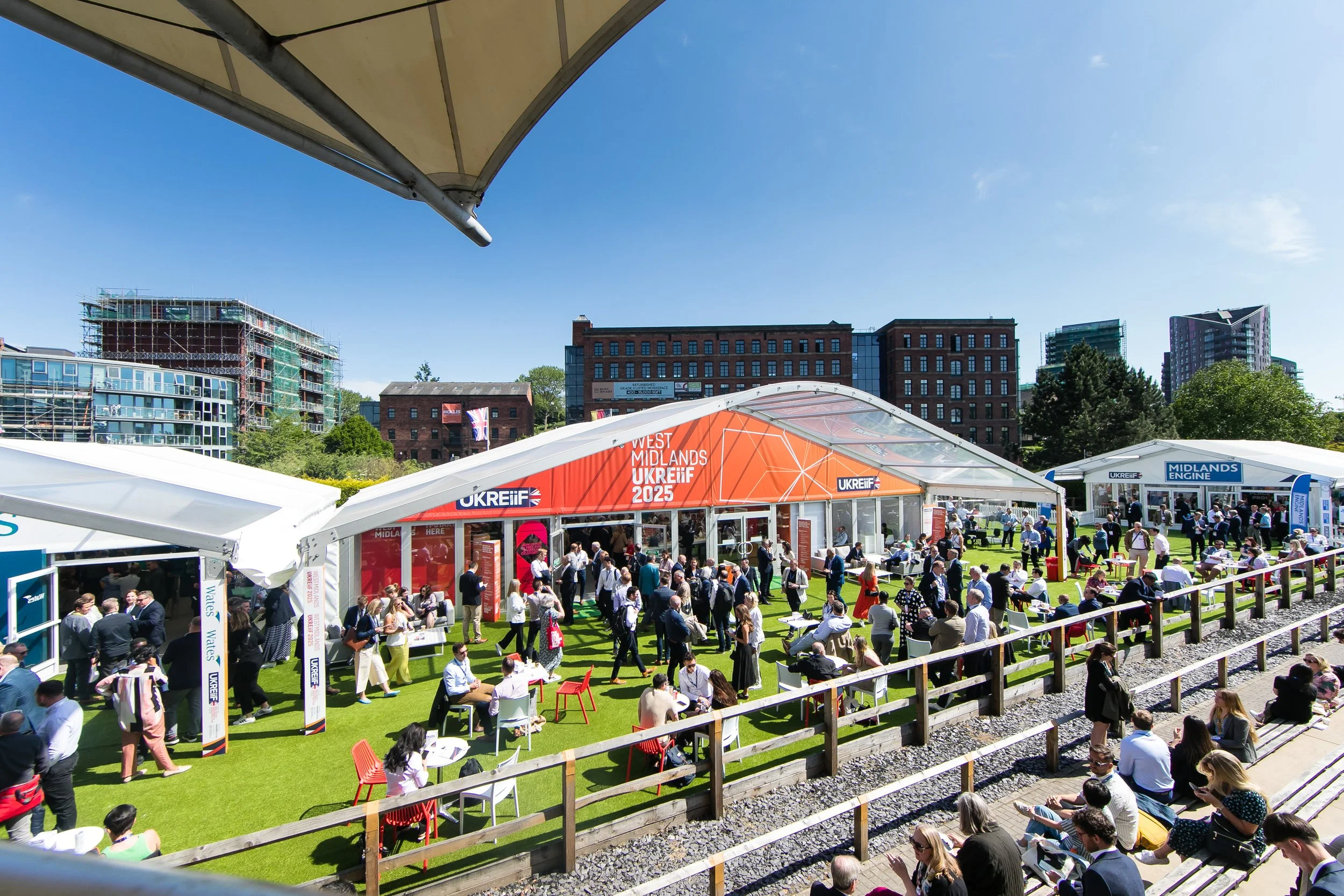 Outdoor event with people gathered under large tents on a sunny day, with some sitting and some walking among the tents, and modern buildings in the background.
