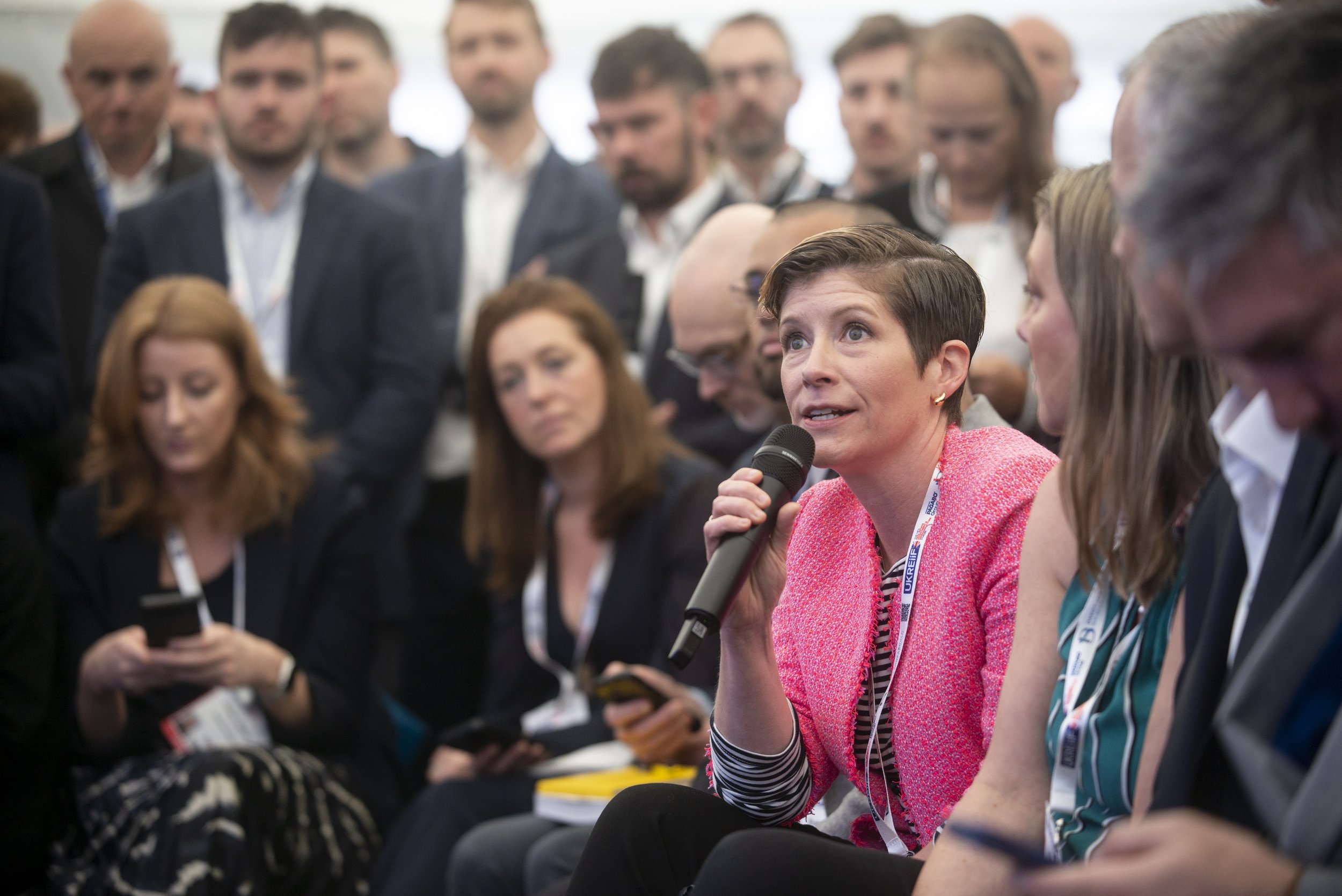 A woman in a pink blazer speaking into a microphone during a panel discussion at a conference, with attentive audience members surrounding her.