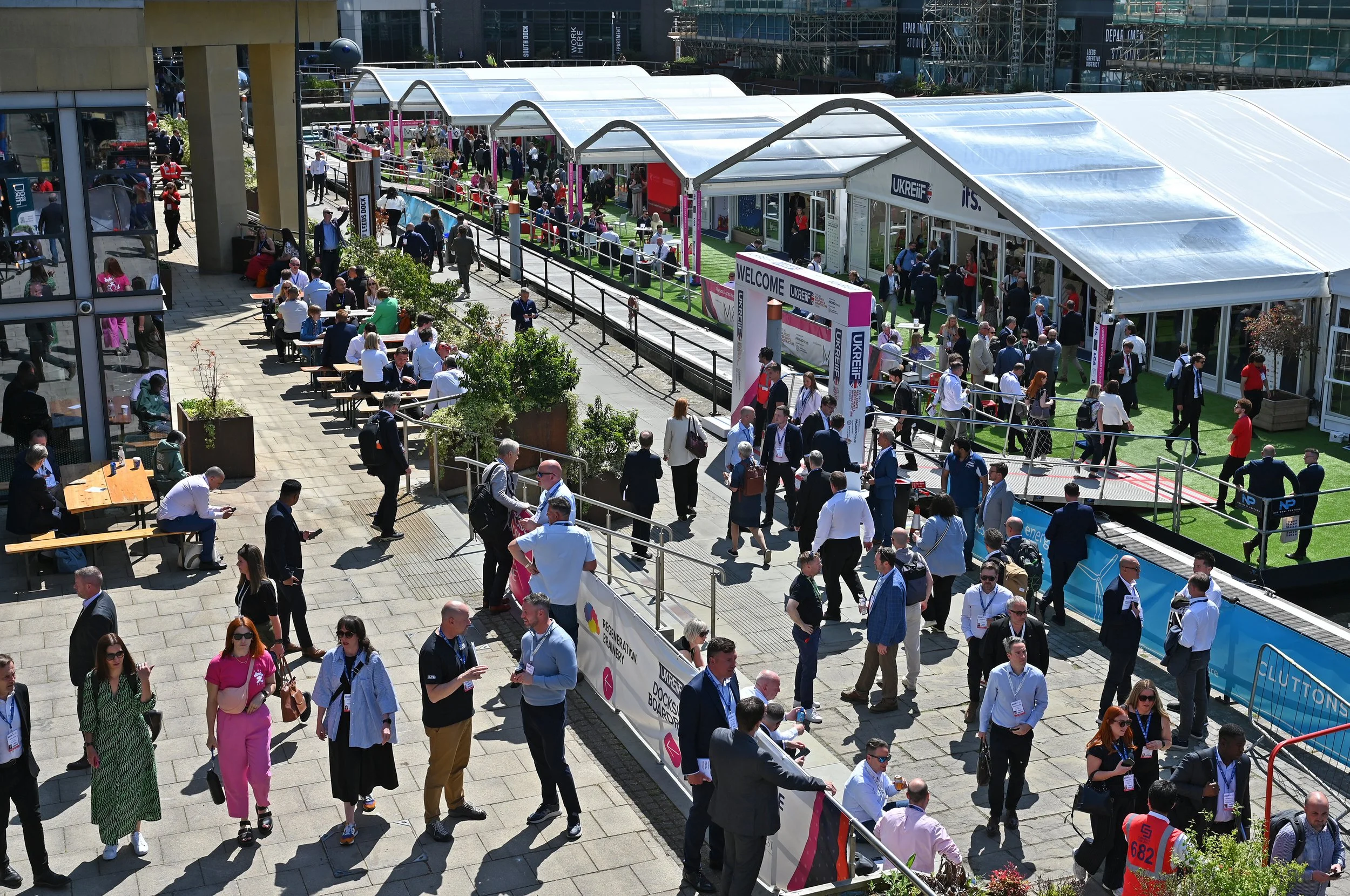 Crowd of people walking and sitting at an outdoor event with booths, tents, and seating areas.