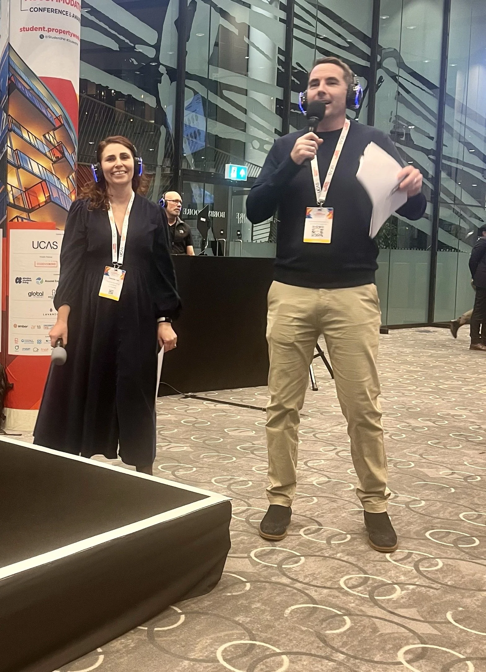 Two people speaking at a conference with a woman holding a microphone and a man with a headset and papers, in a large glass-walled conference hall.