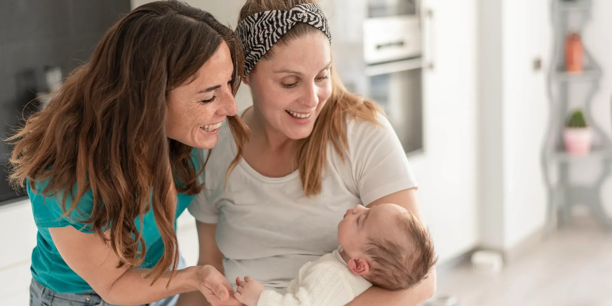 Two women and a baby smiling and looking at each other in a kitchen.