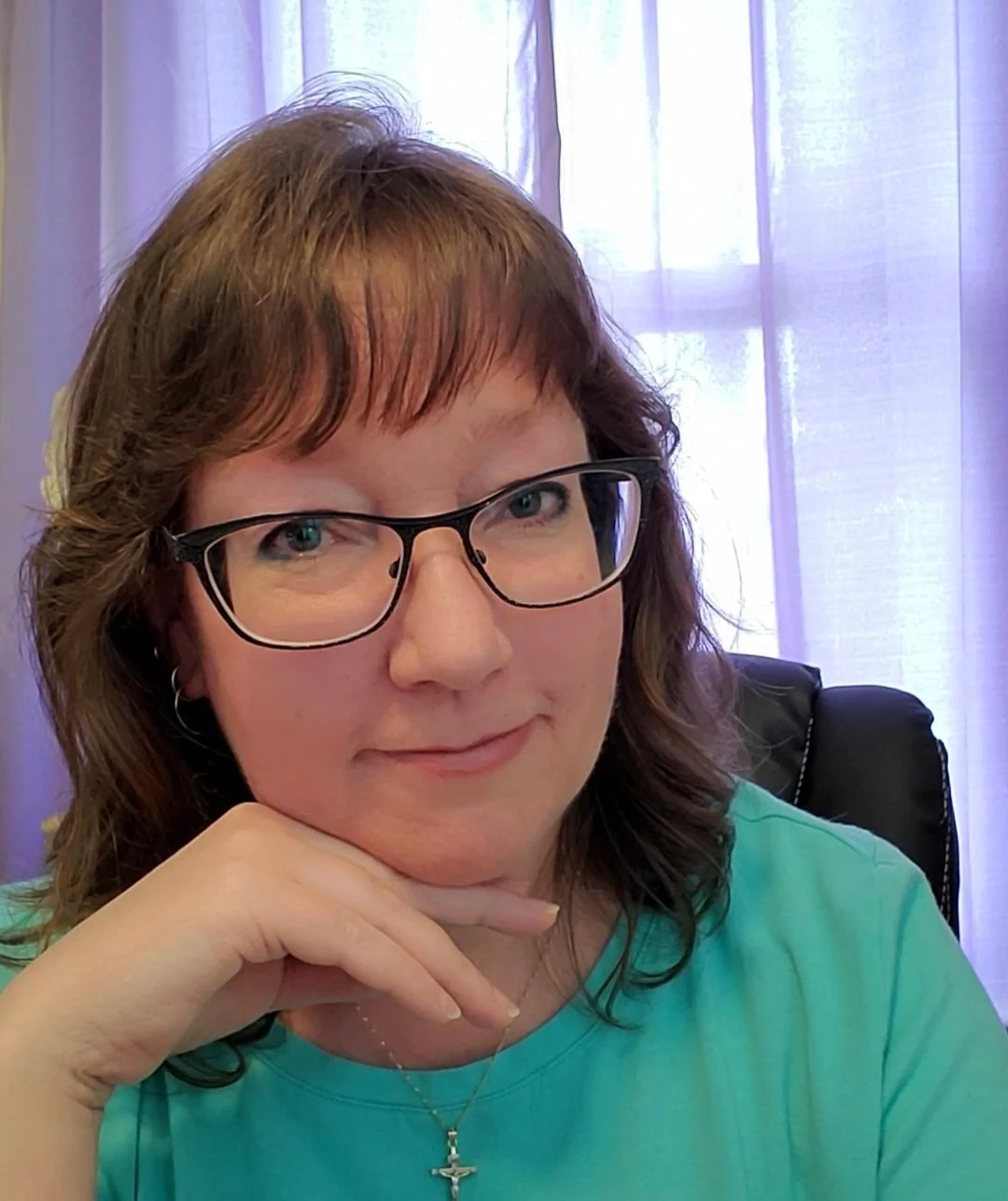A woman with shoulder-length brown hair wearing glasses, a turquoise shirt, a cross necklace, and a small hoop earring, sitting in front of purple curtains.