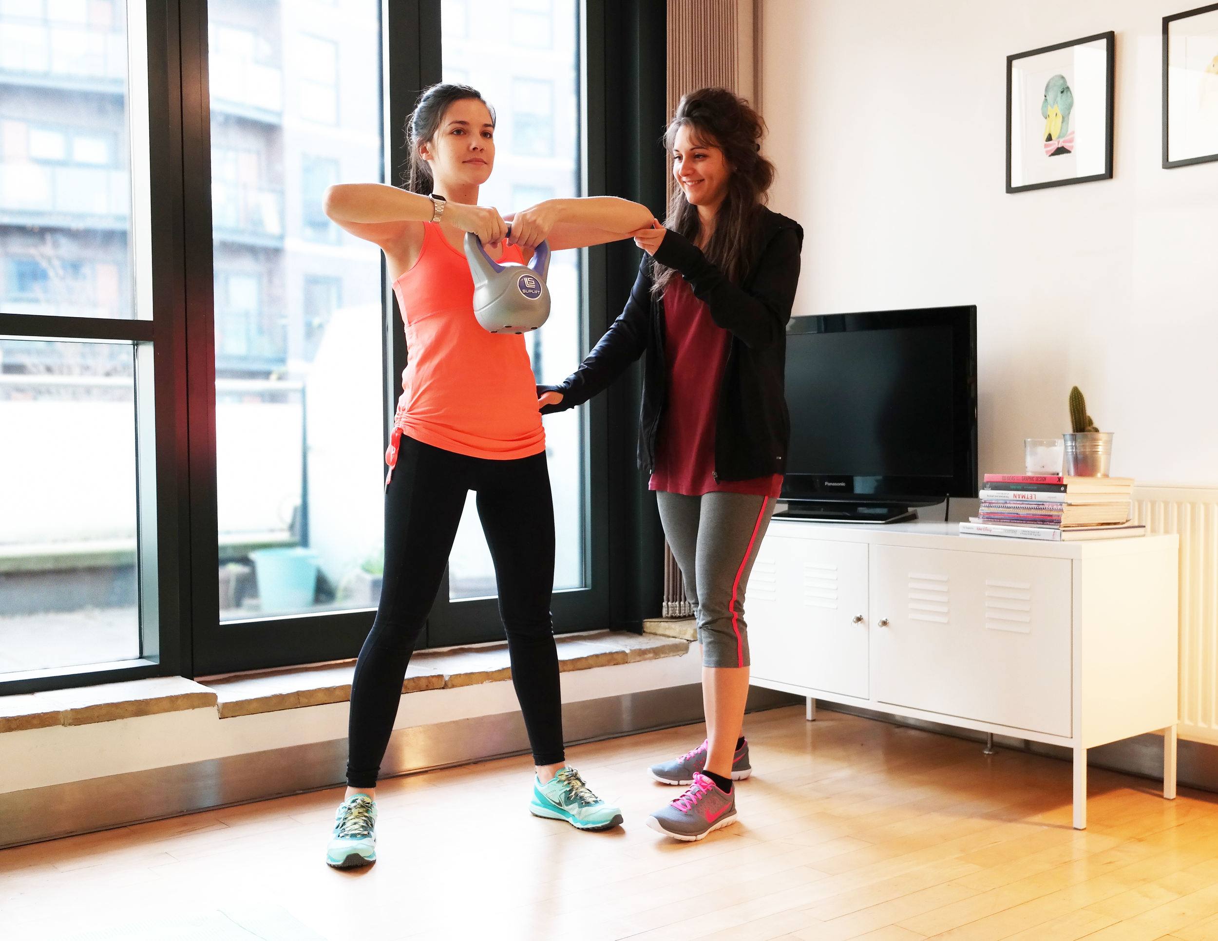 Two women in workout clothes in a living room with a large window. One woman is holding a kettlebell, and the other woman is assisting or coaching.