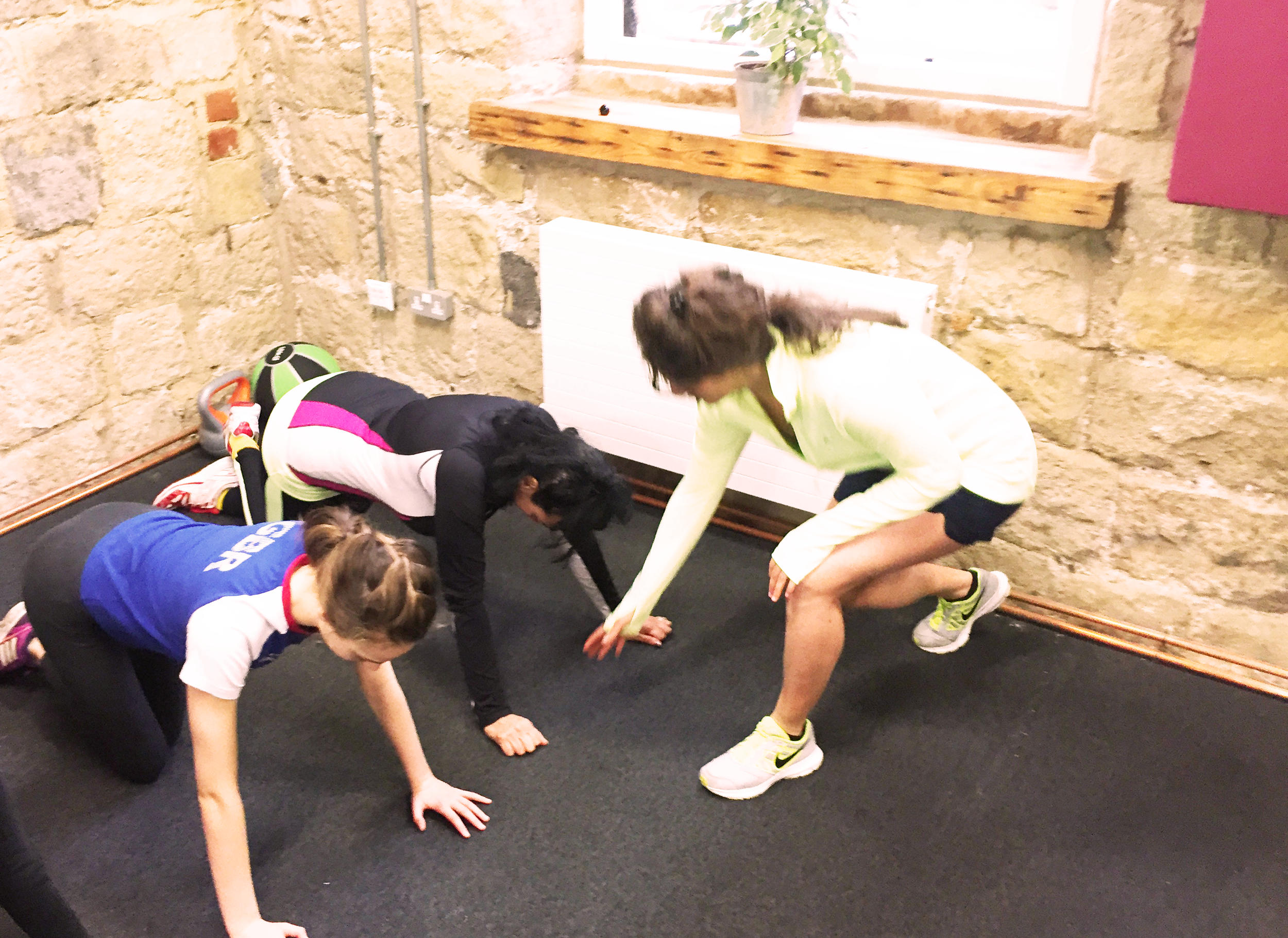 Three women doing a planking exercise on their hands and knees on a black mat in a room with exposed brick walls and a window with a plant on the sill.