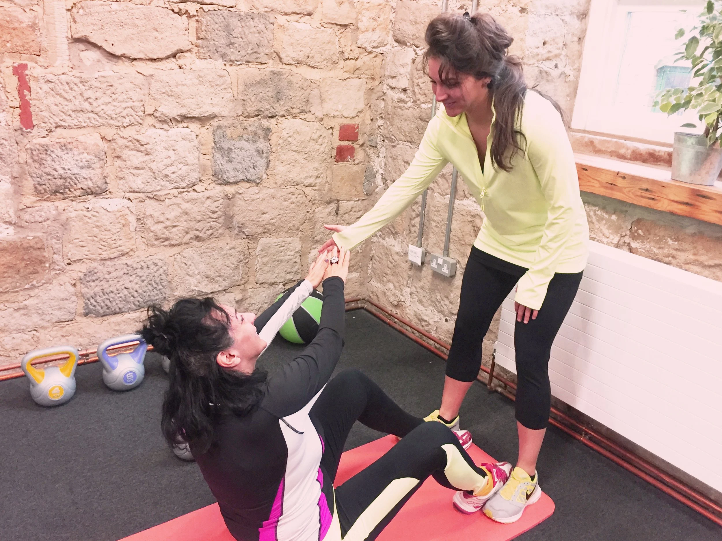 A woman in black and pink workout clothes sitting on a pink exercise mat helping another woman in yellow workout clothes with stethoscope around her neck during fitness training in a brick-walled gym.
